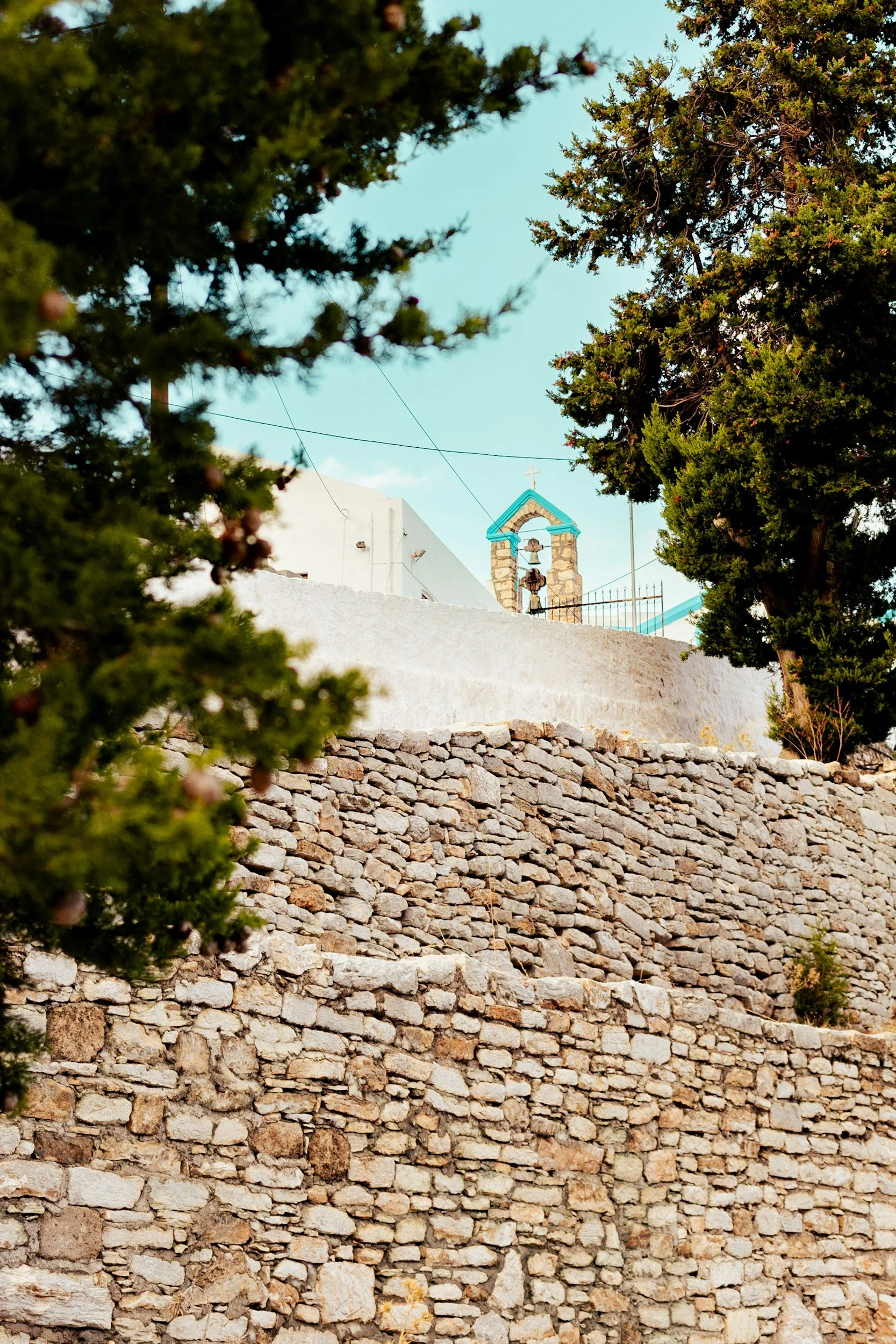 A stone wall with a small white church building and a bell tower with a blue roof visible above, framed by trees not far from The Selene Greece in Symi.