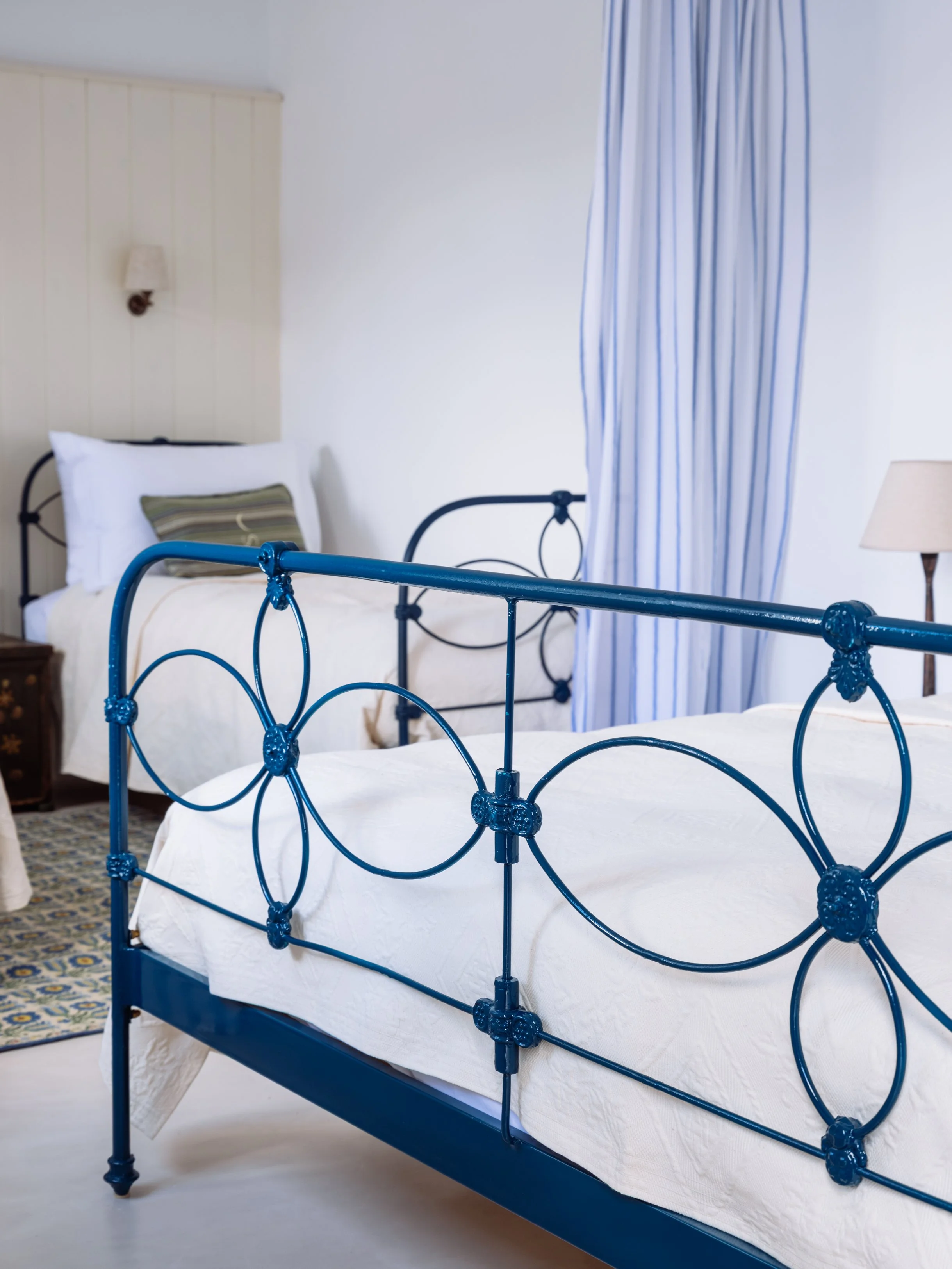 Gaia suite with antique blue metal bed frame, white bedding, pillow, wooden nightstand, wall-mounted lamp, and blue and white striped curtains at the selene greece, symi