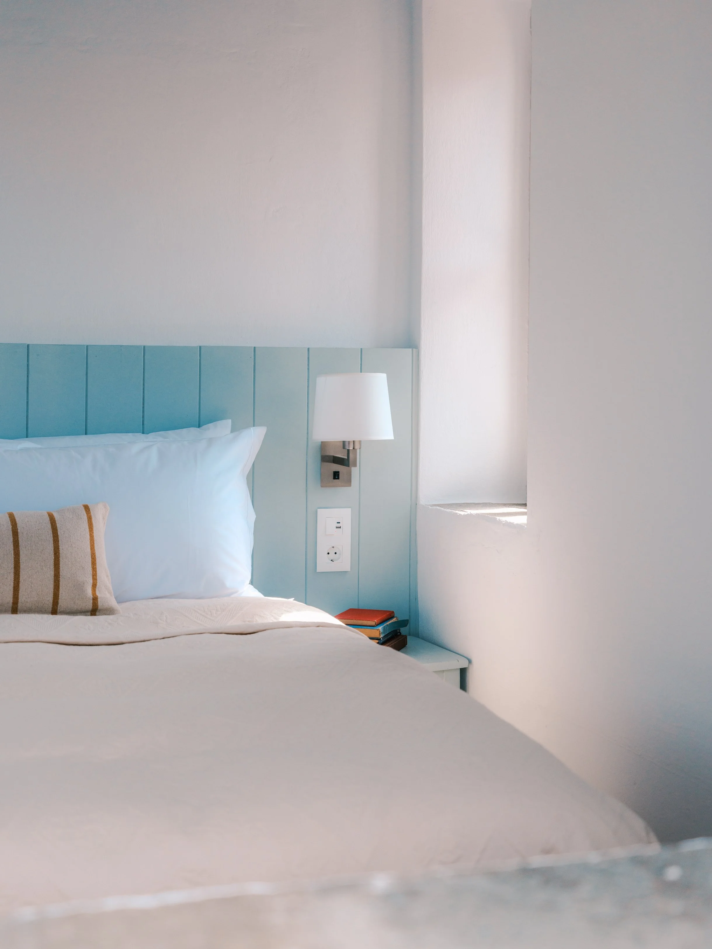 Eos room king bed with white bedding and pillows. The bed has a light blue headboard, with a mounted bedside lamp and a small stack of books on the nightstand.  At The Selene Greece, Symi