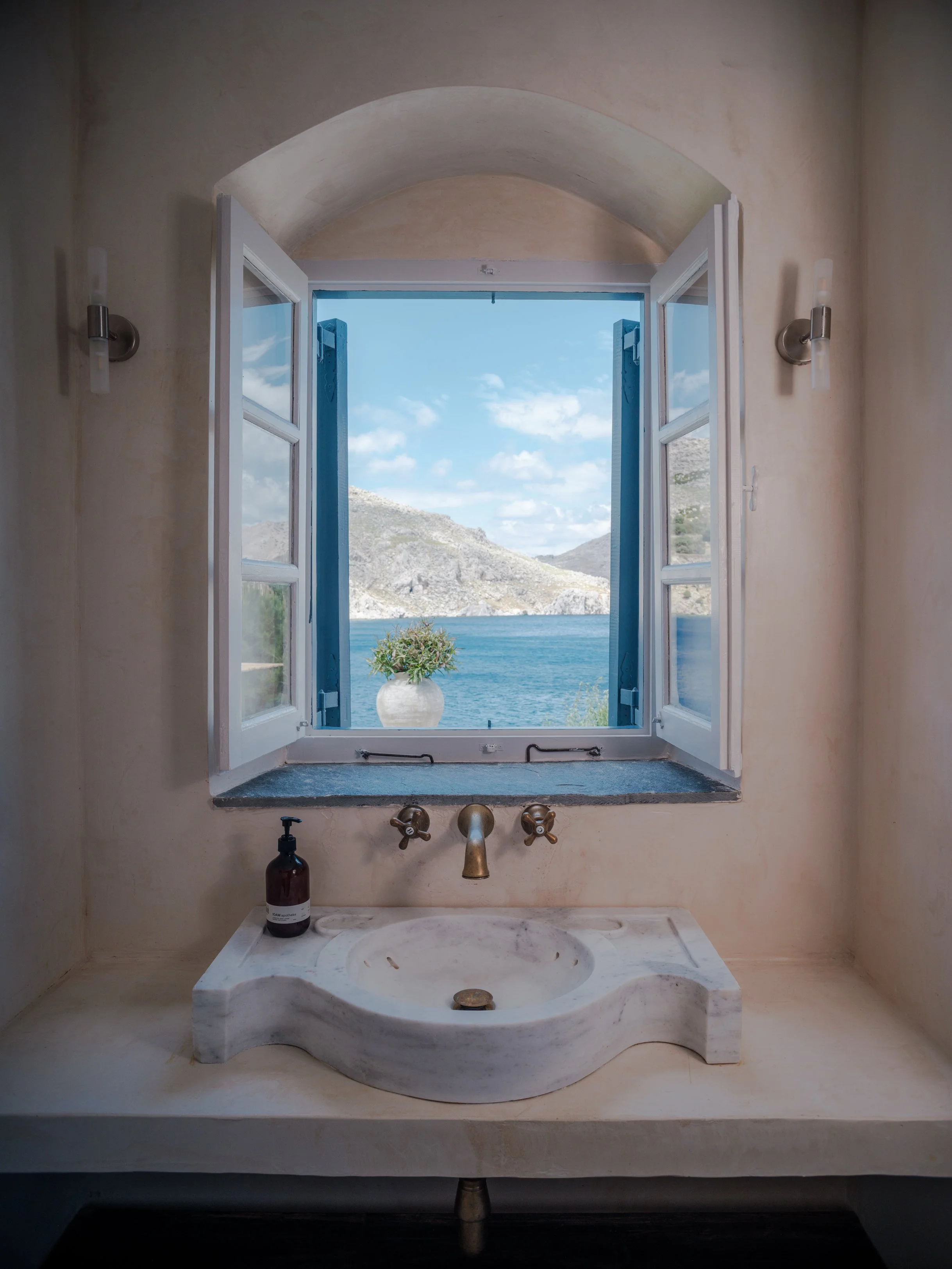 Gaia suite bathroom sink with a marble basin, two vintage-style brass fixtures, and an open window showing a scenic view of nimborio bay, mountains and a blue sky at the selene greece, symi