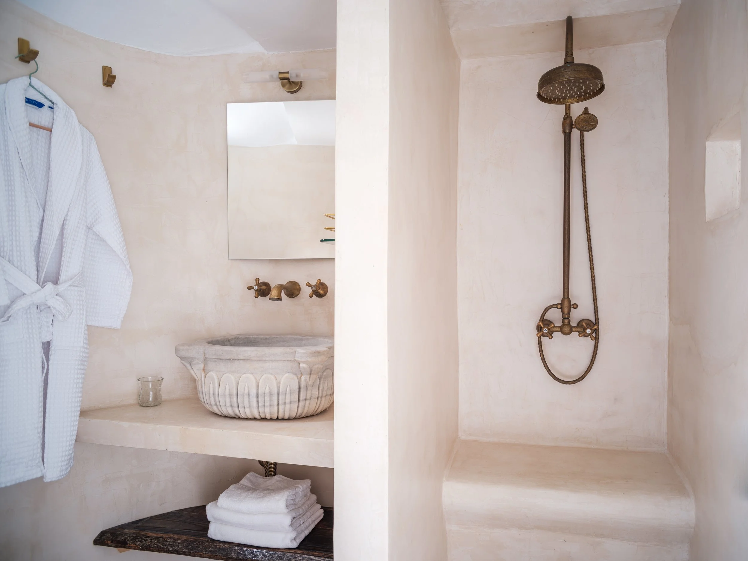 Eos room bathroom with a brass shower, a small mirror, and a white bathrobe hanging on a wall hook. A marble sink is set into a shelf with stacked white towels underneath. At The Selene Greece, Symi