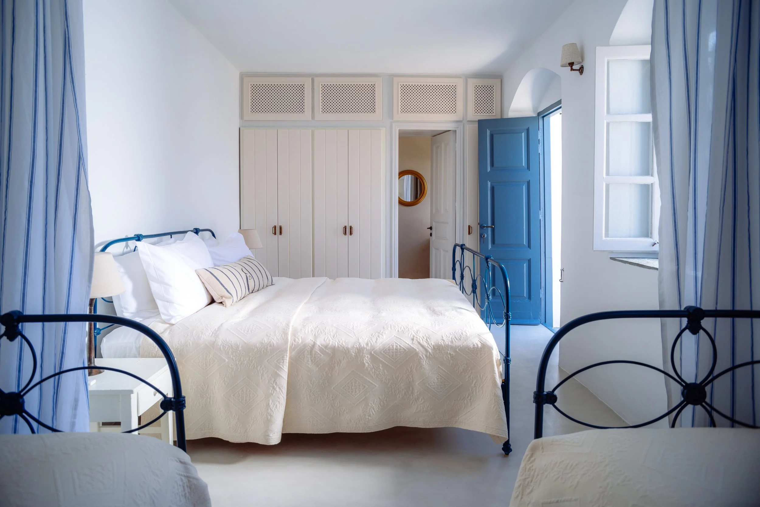 Gaia suite showing king size bed and a striped pillow, blue door and matching blue curtains, and a window with sunlight coming through at the selene greece, symi