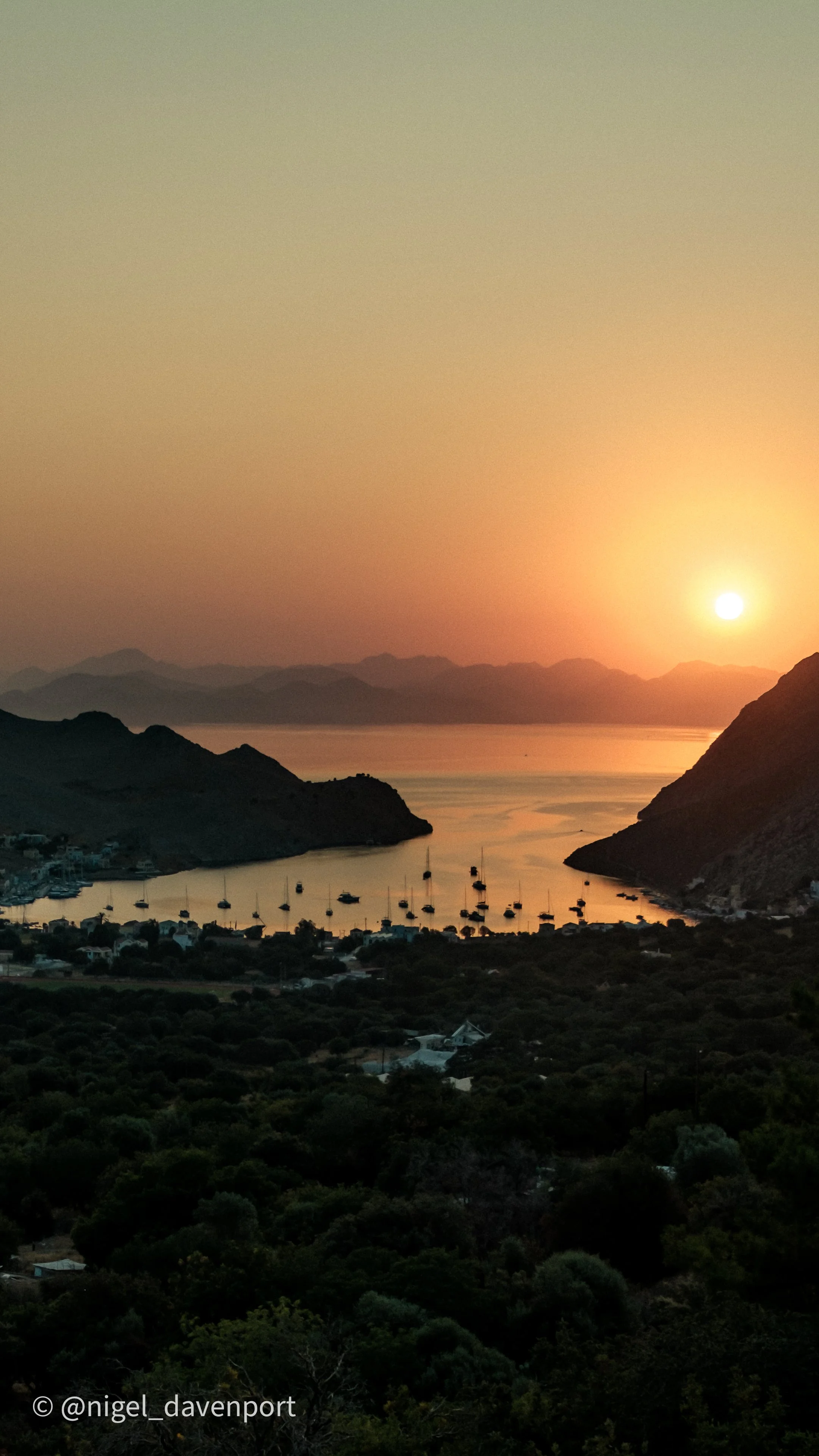 Sunset over Pedi bay in Symui Greece, with boats anchored, surrounded by mountains and lush greenery.