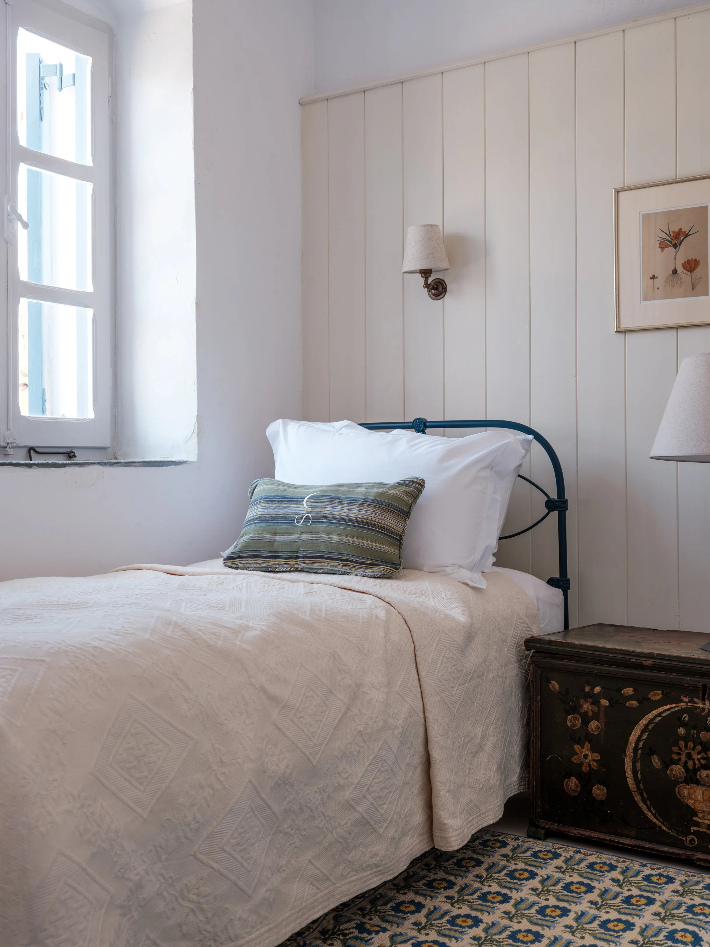 Gaia suite single bed with bedspread, a pillow with a striped pillowcase, a window letting in natural light, a wall-mounted lamp, a framed floral artwork, a vintage wooden nightstand with floral designs, and a patterned rug at the selene greece, symi