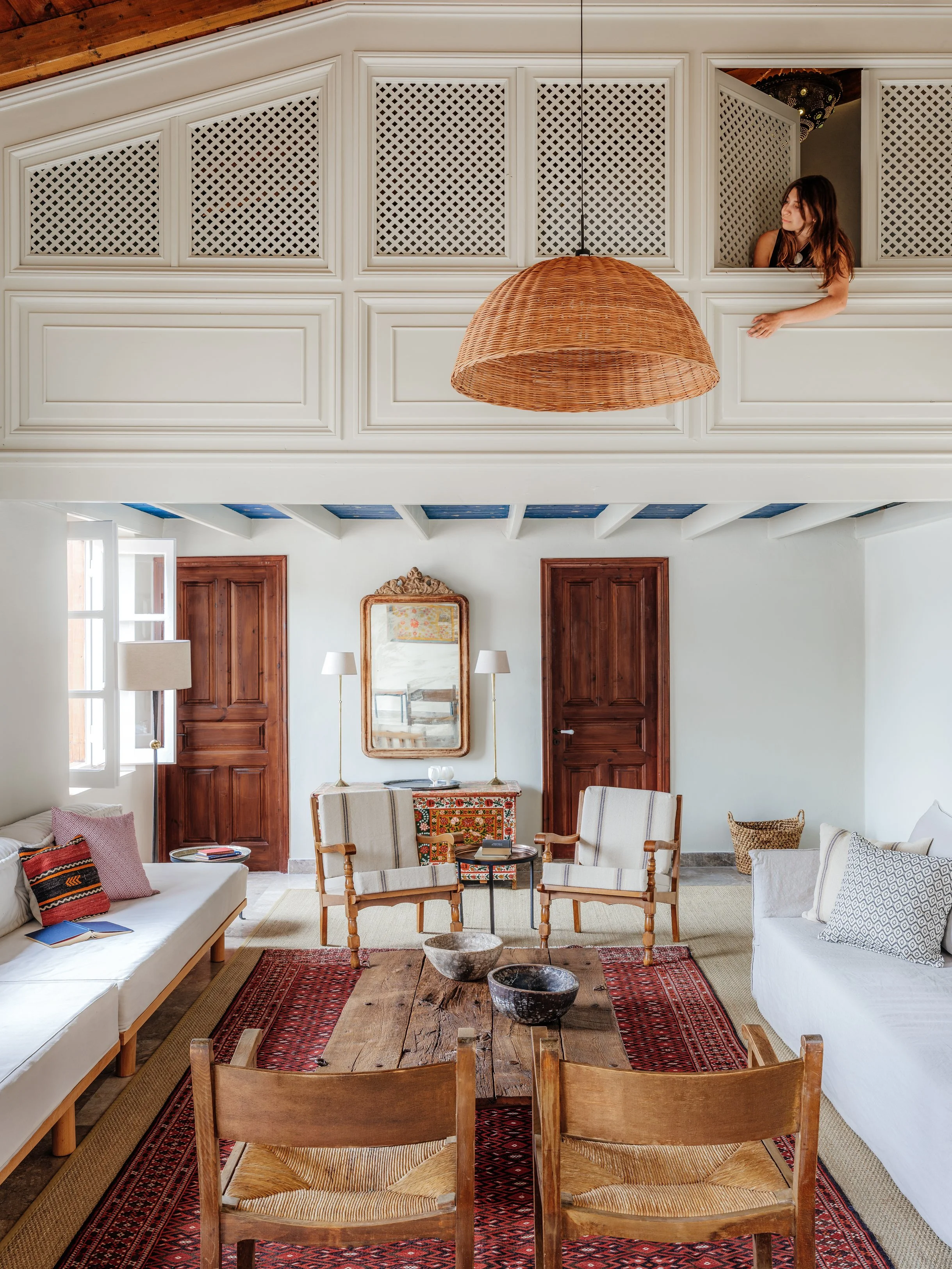 Living room with two sofas. A girl peeks out from a balcony above, which has white lattice panels, and a large wicker lampshade hanging from the ceiling. At The Selene Greece, Symi
