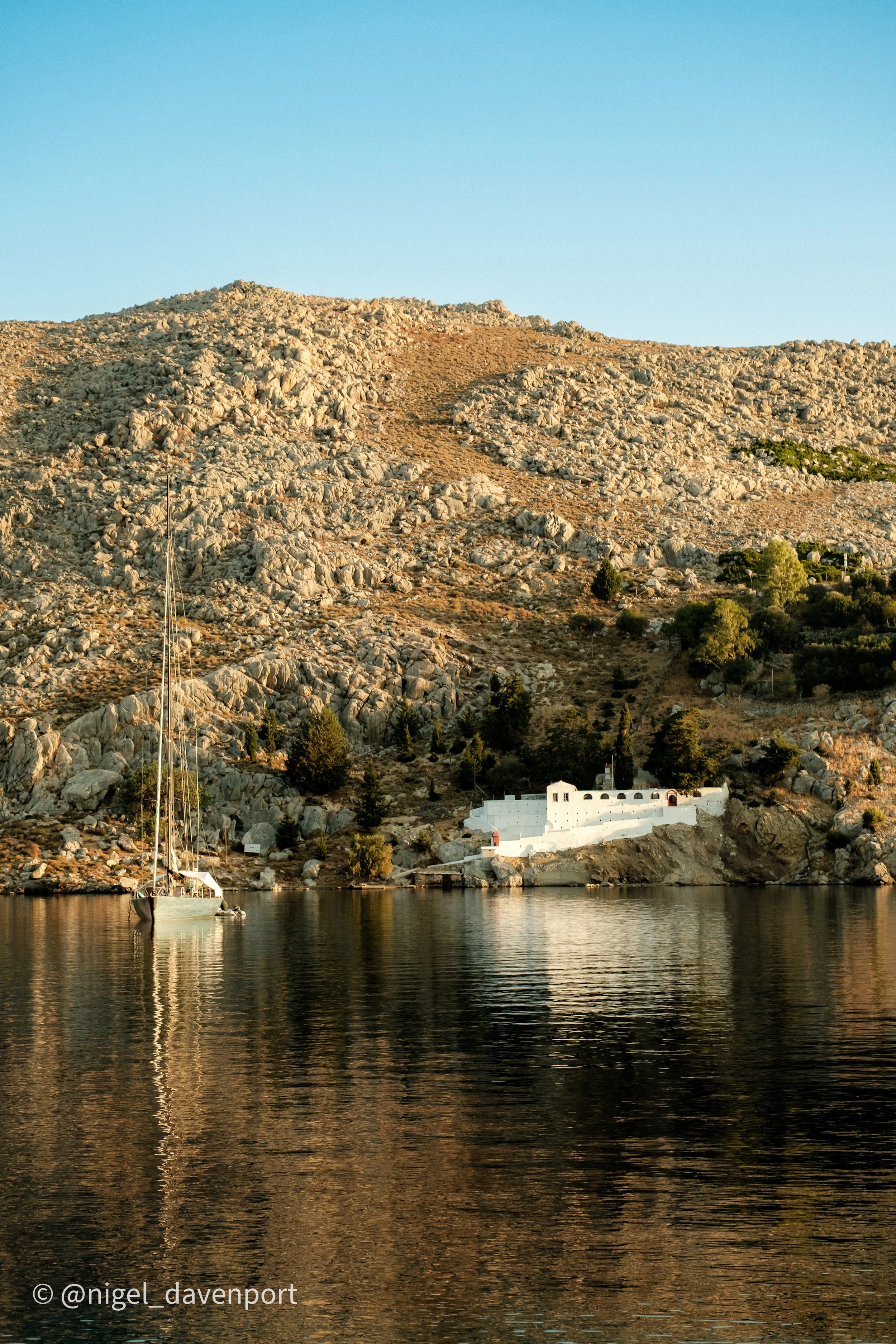 A view from The Selene Greece out over Nimborio Bay in Symi. A sailboat anchored near rocky shore with the monastery in the distance, and trees at the base of a mountain