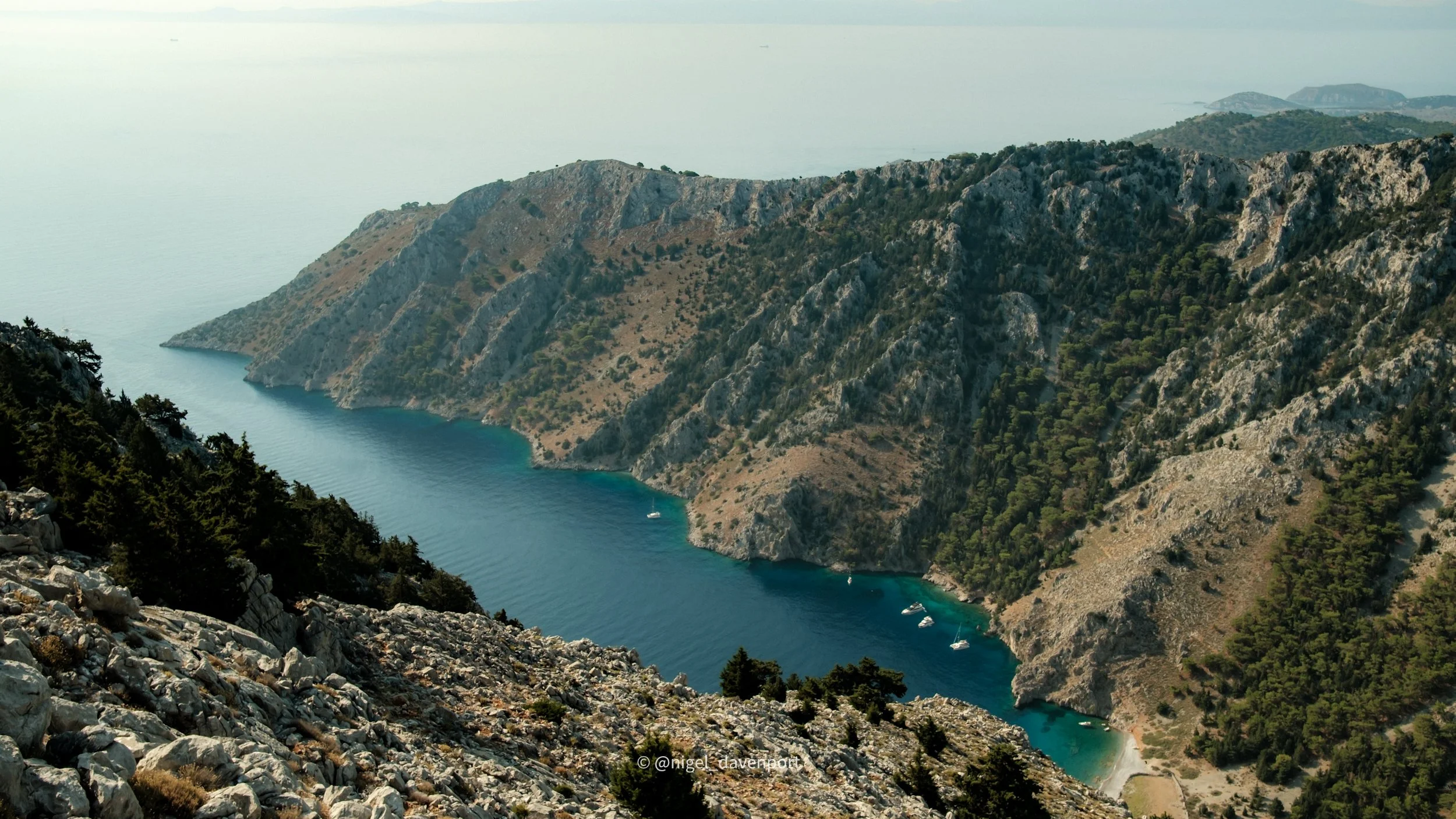 A scenic view of a coastal landscape with steep rocky cliffs covered in greenery, looking down on Nanou bay in Symi Greece