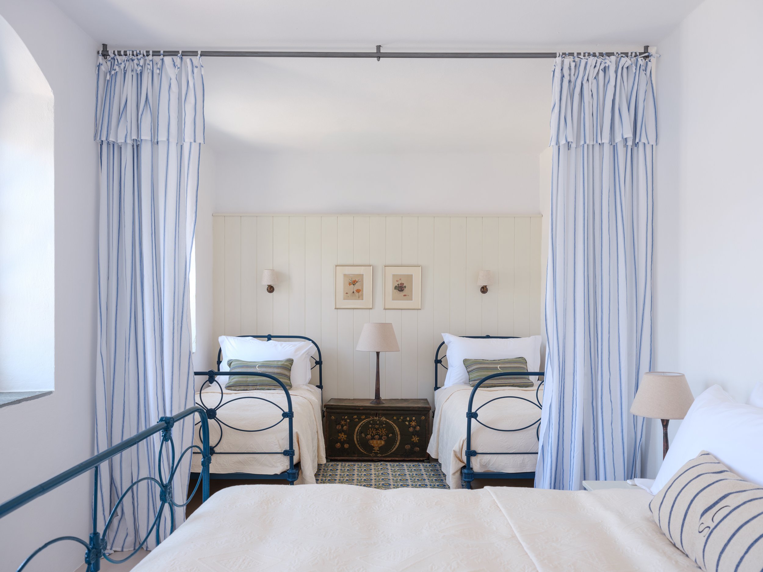 Gaia bedroom with two iron beds, blue and white striped curtains, framed floral artwork, beige lamps, and a narrow dark chest at the foot of the beds, in a bright room with white walls at the selene greece, symi
