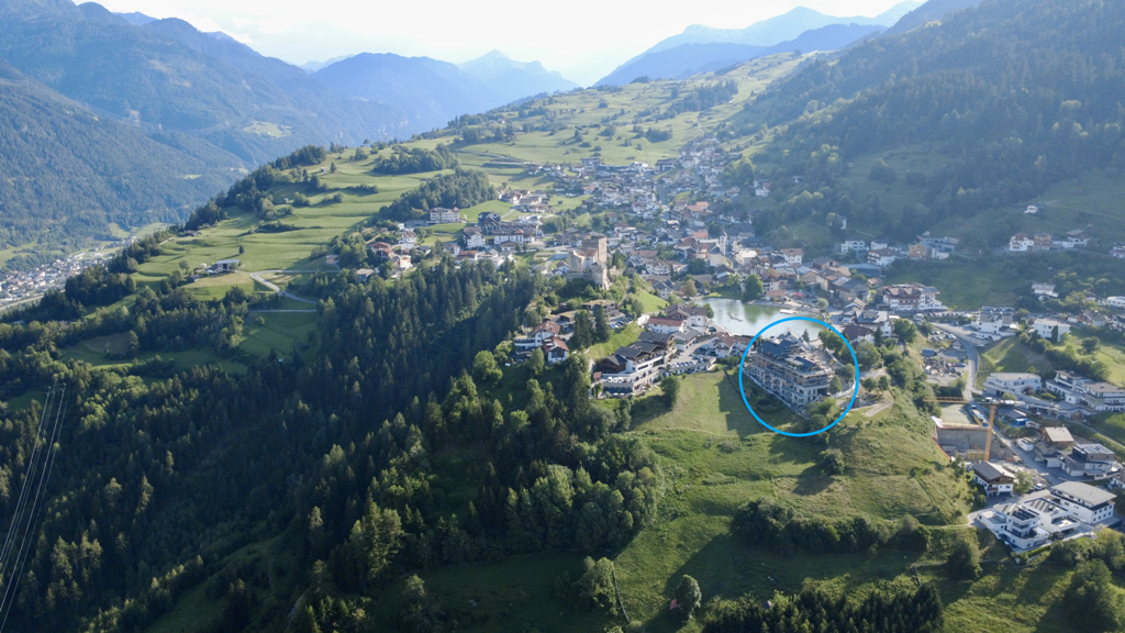 Luftaufnahme der bergigen Landschaft um Ladis und den grünen Wiese, die von Bäumen gesäumt sind. In der Mitte rechts ist die homelodge mit einem blauen Kreis markiert.