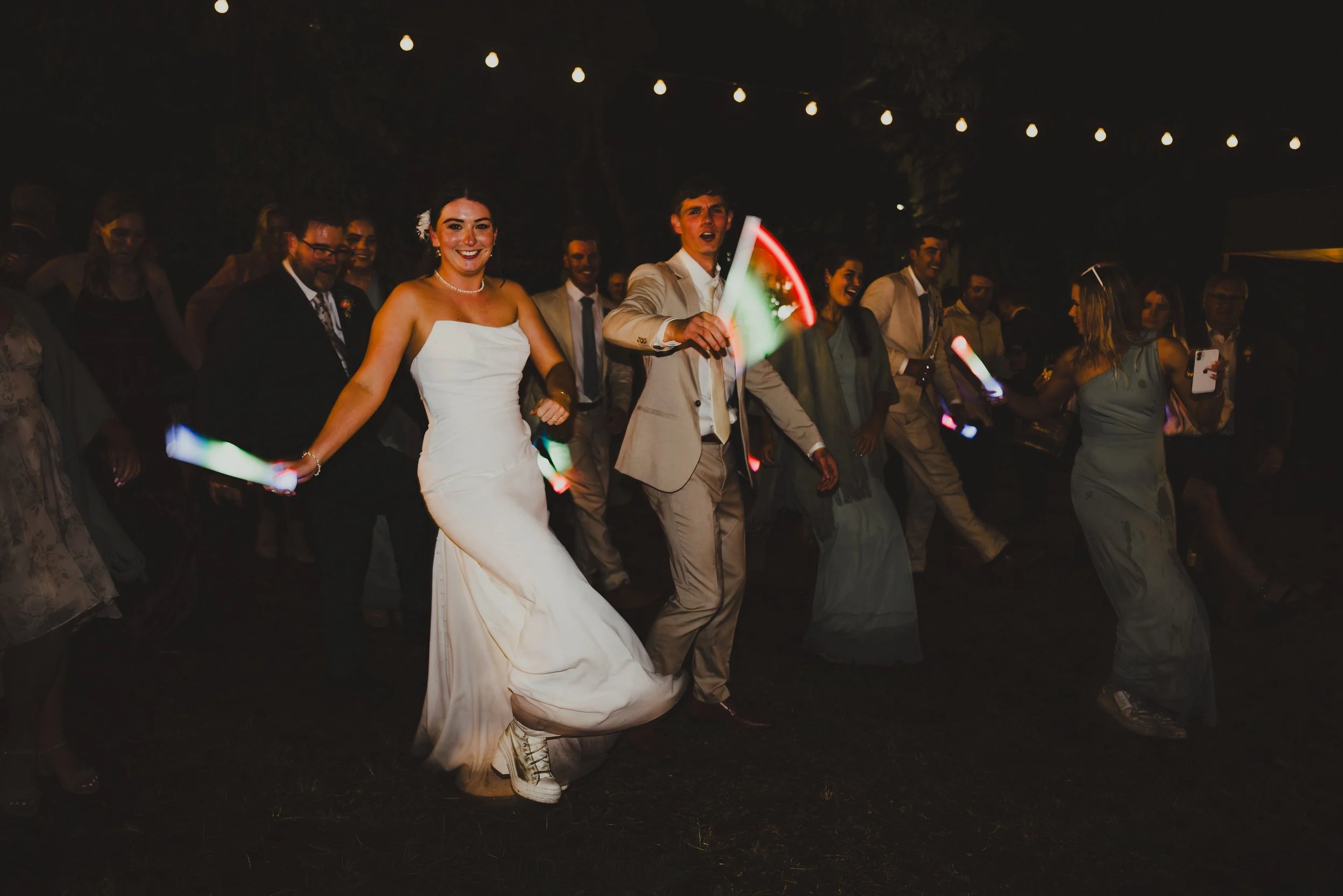 People dancing at a nighttime celebration, including a woman in a white wedding dress and a man in a beige suit holding glow sticks, with string lights overhead.