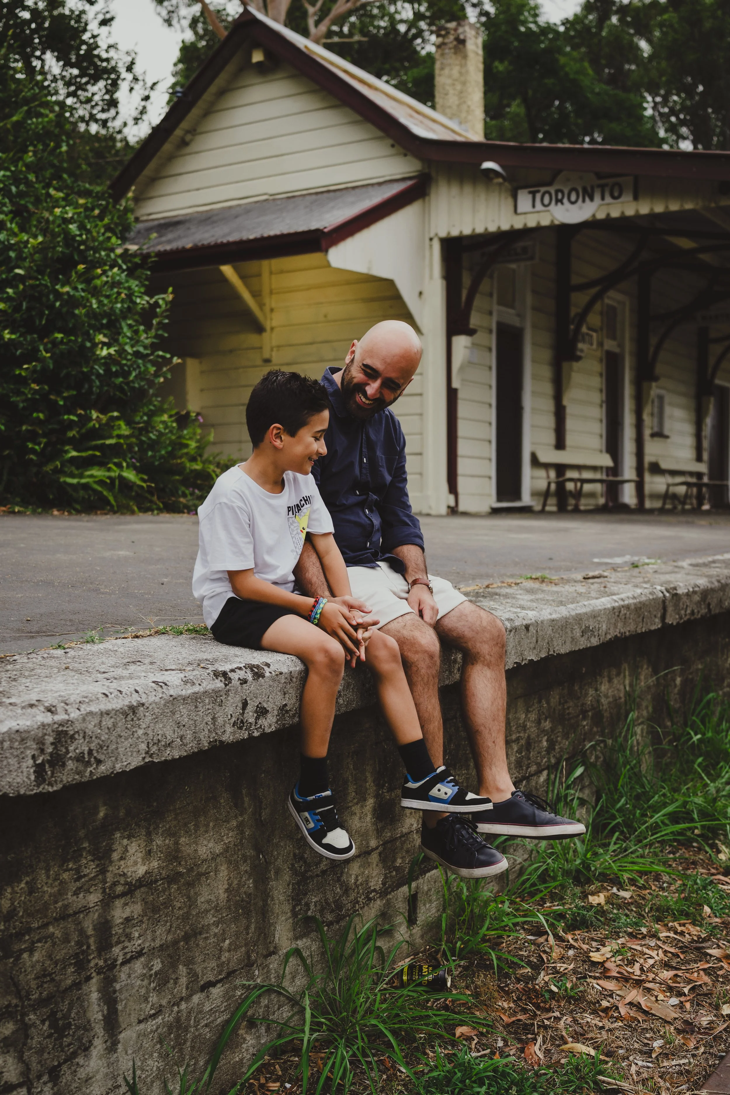 A man and a boy sitting on a concrete ledge at a train station, smiling and laughing together.