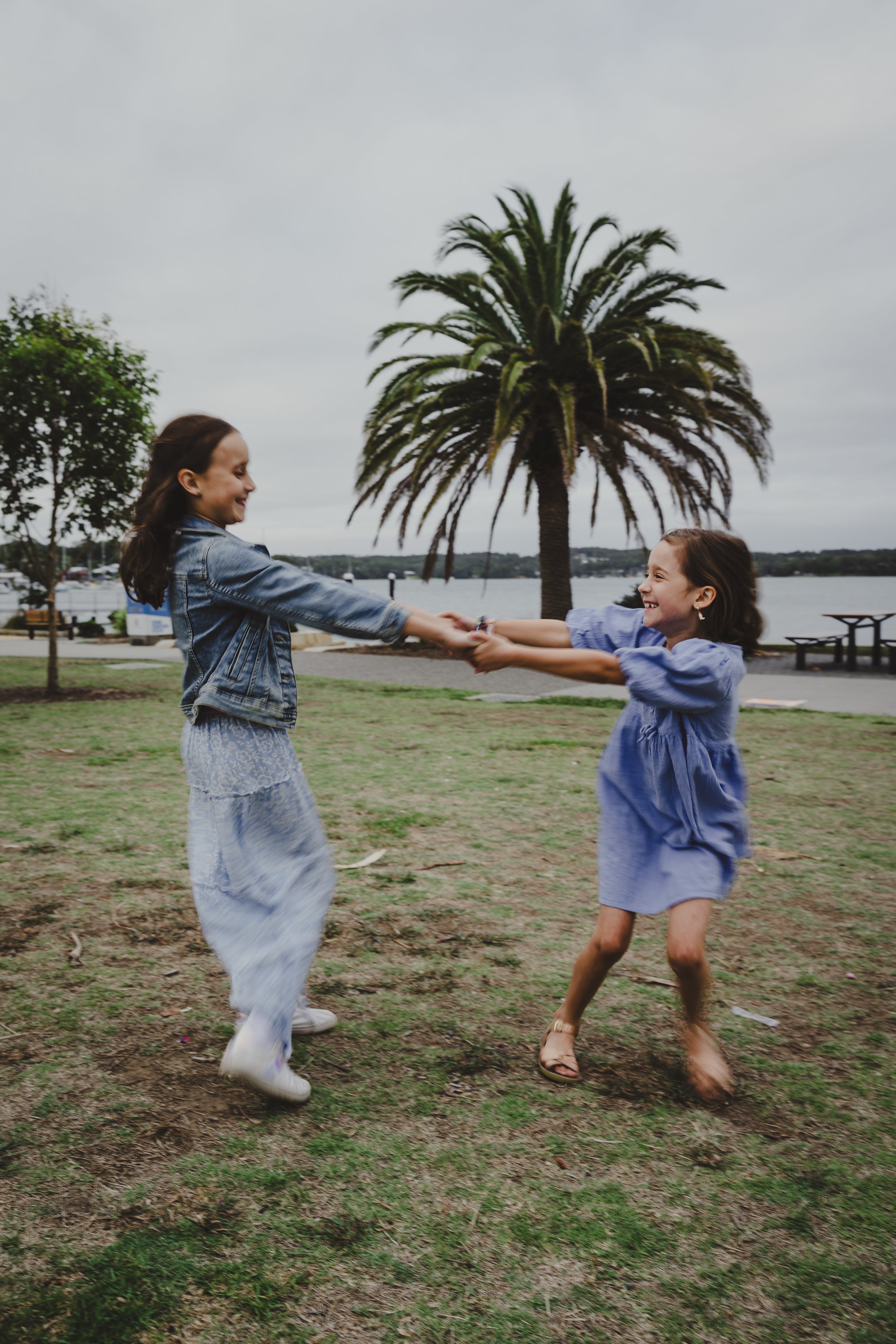 Two girls, one in a denim jacket and the other in a blue dress, are playing and laughing in a park with a large palm tree and water in the background.