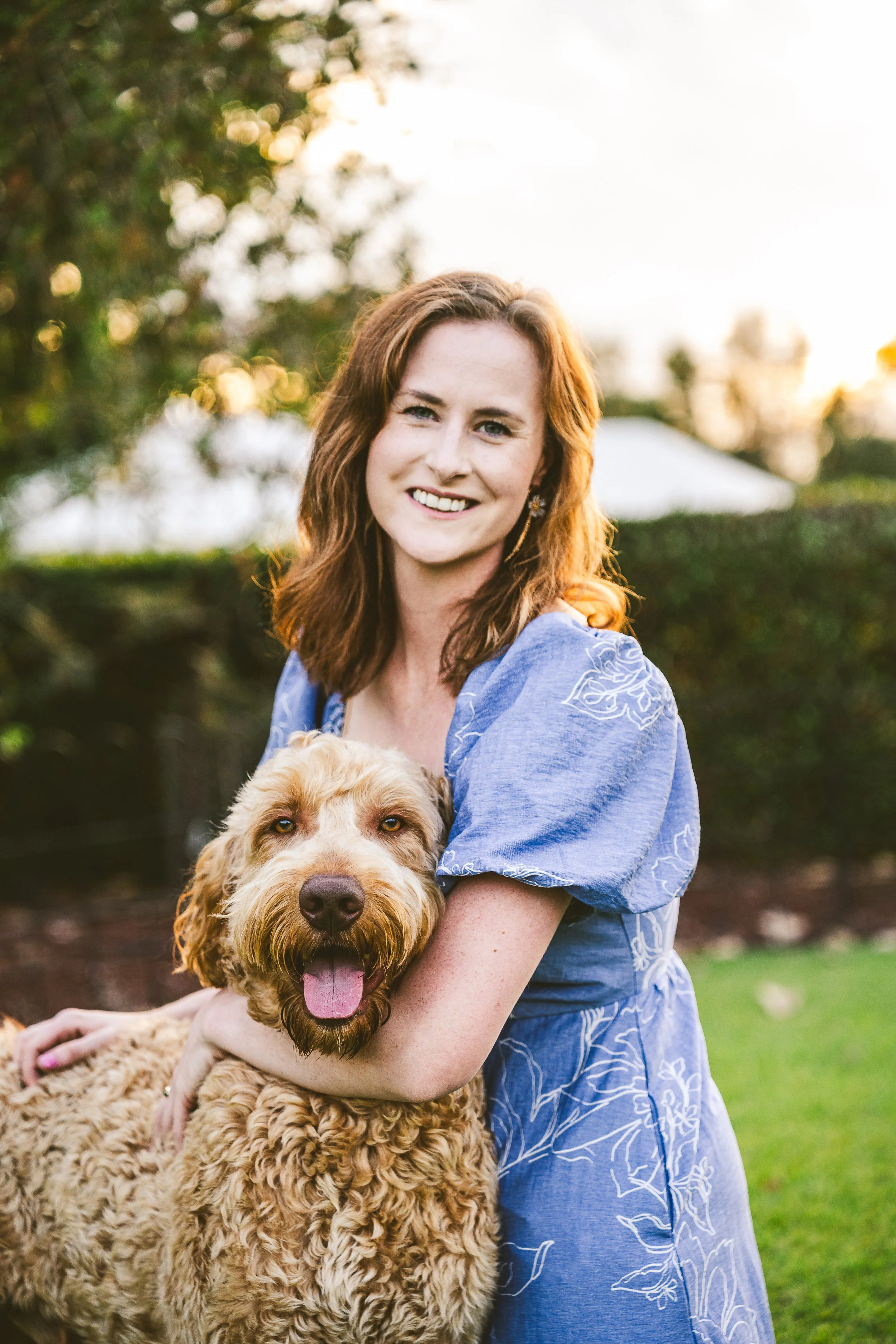 A woman with red hair smiling while holding a large, curly-haired golden retriever dog outdoors in a park during golden hour.
