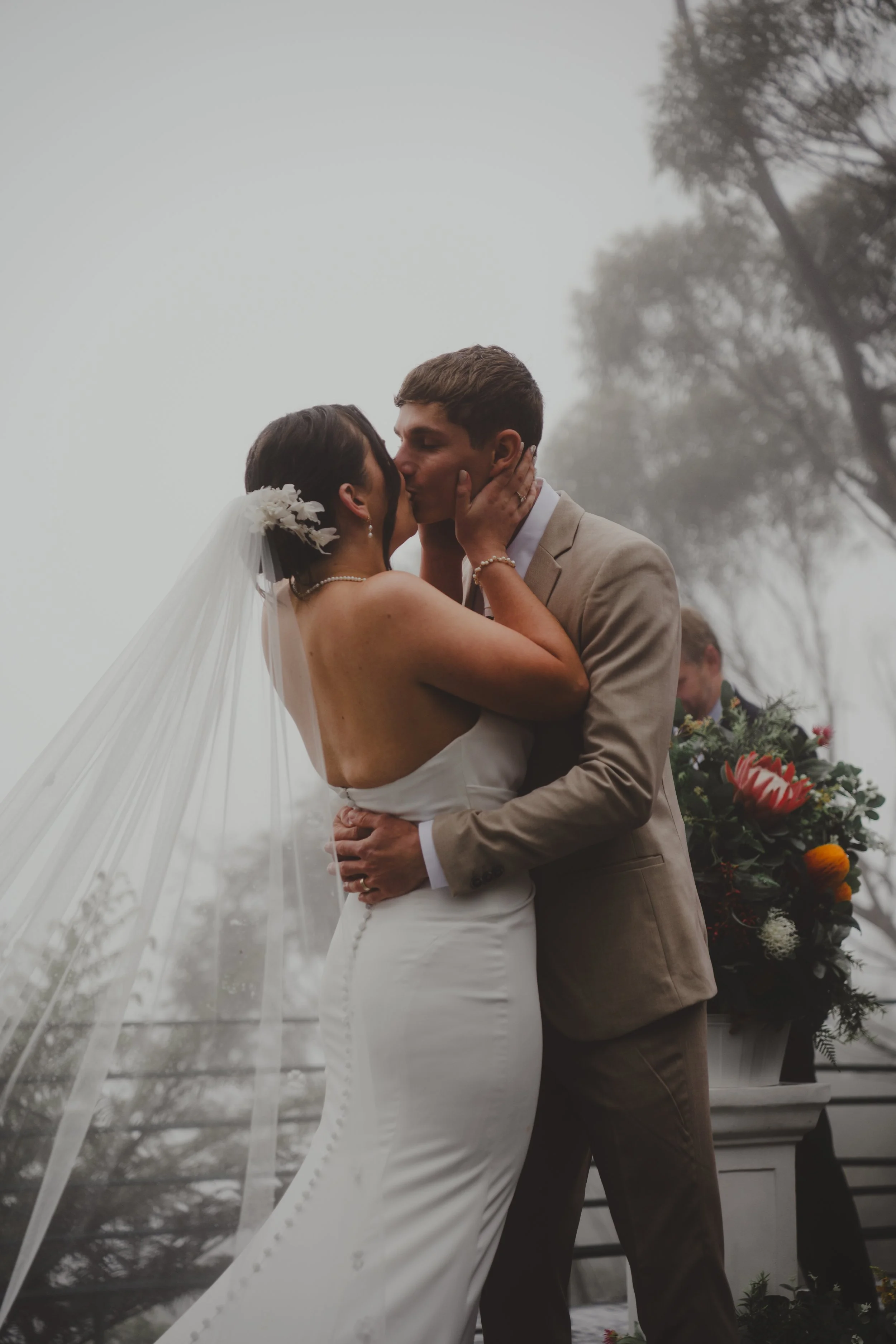 Bride and groom kissing outdoors on their wedding day, foggy weather with trees in the background, a large floral arrangement nearby.