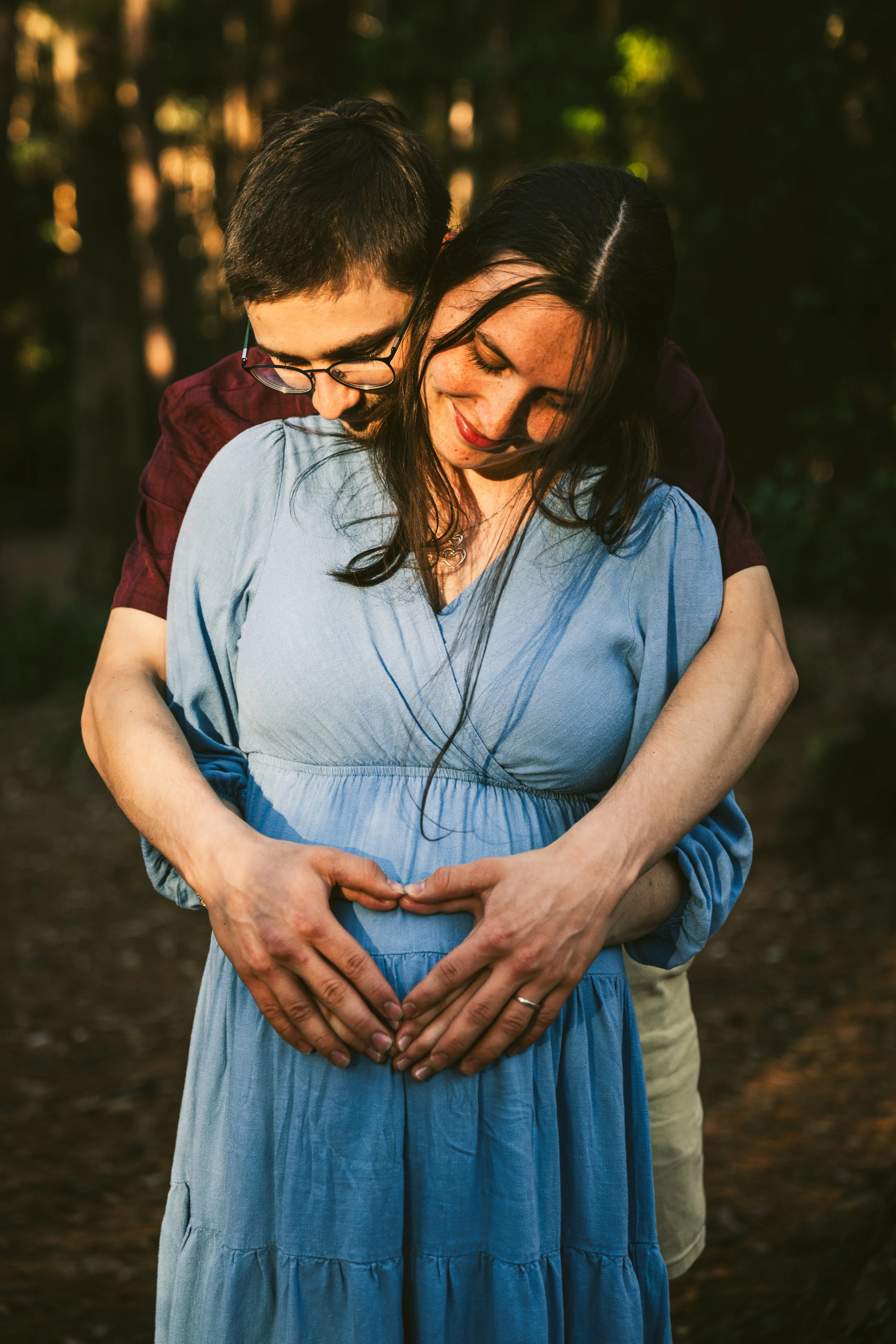 A couple standing outdoors in a forest, with the man embracing the woman from behind as they make a heart shape with their hands on her pregnant belly.