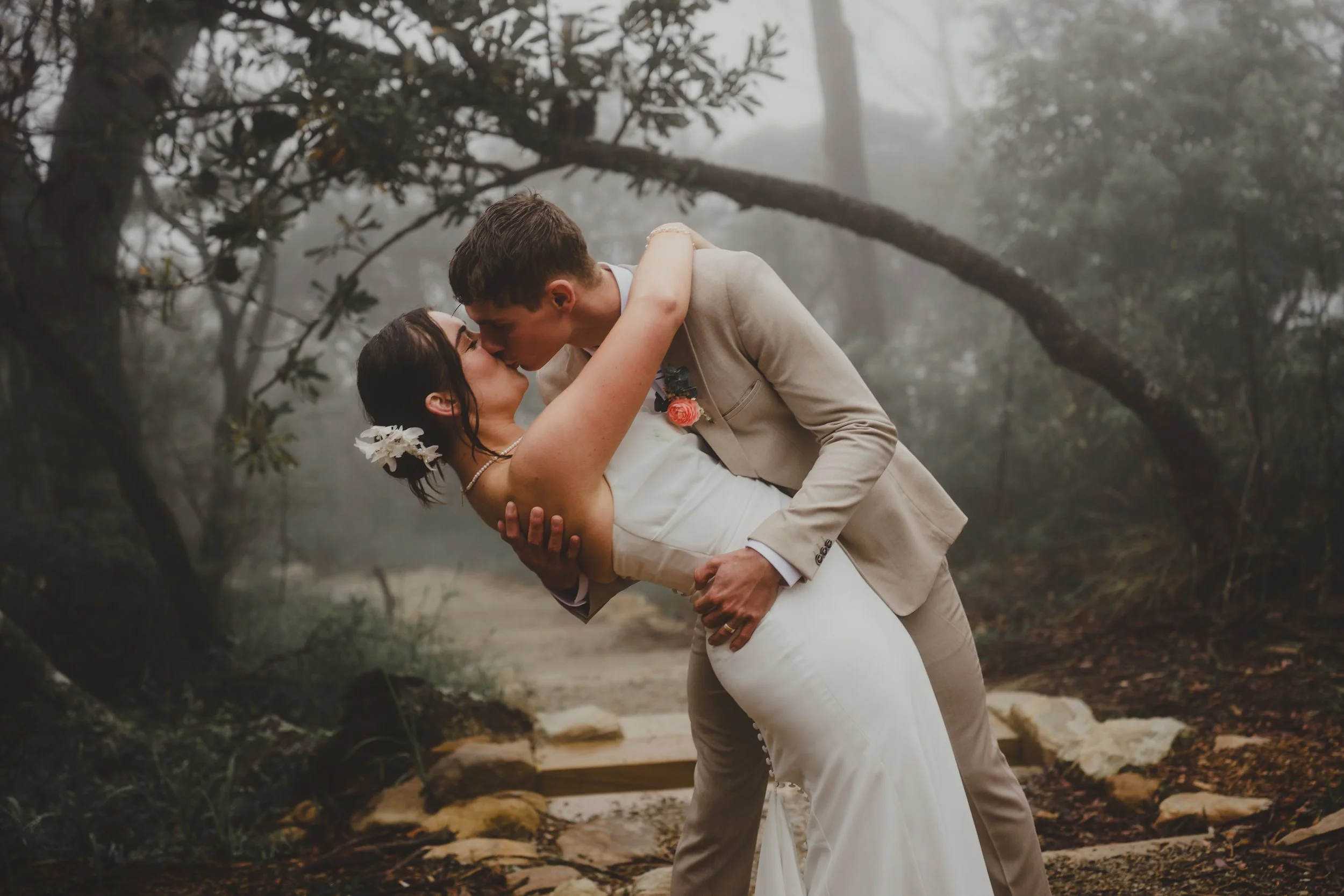 A couple in wedding attire sharing a kiss in a foggy forest, with the groom dipping the bride.