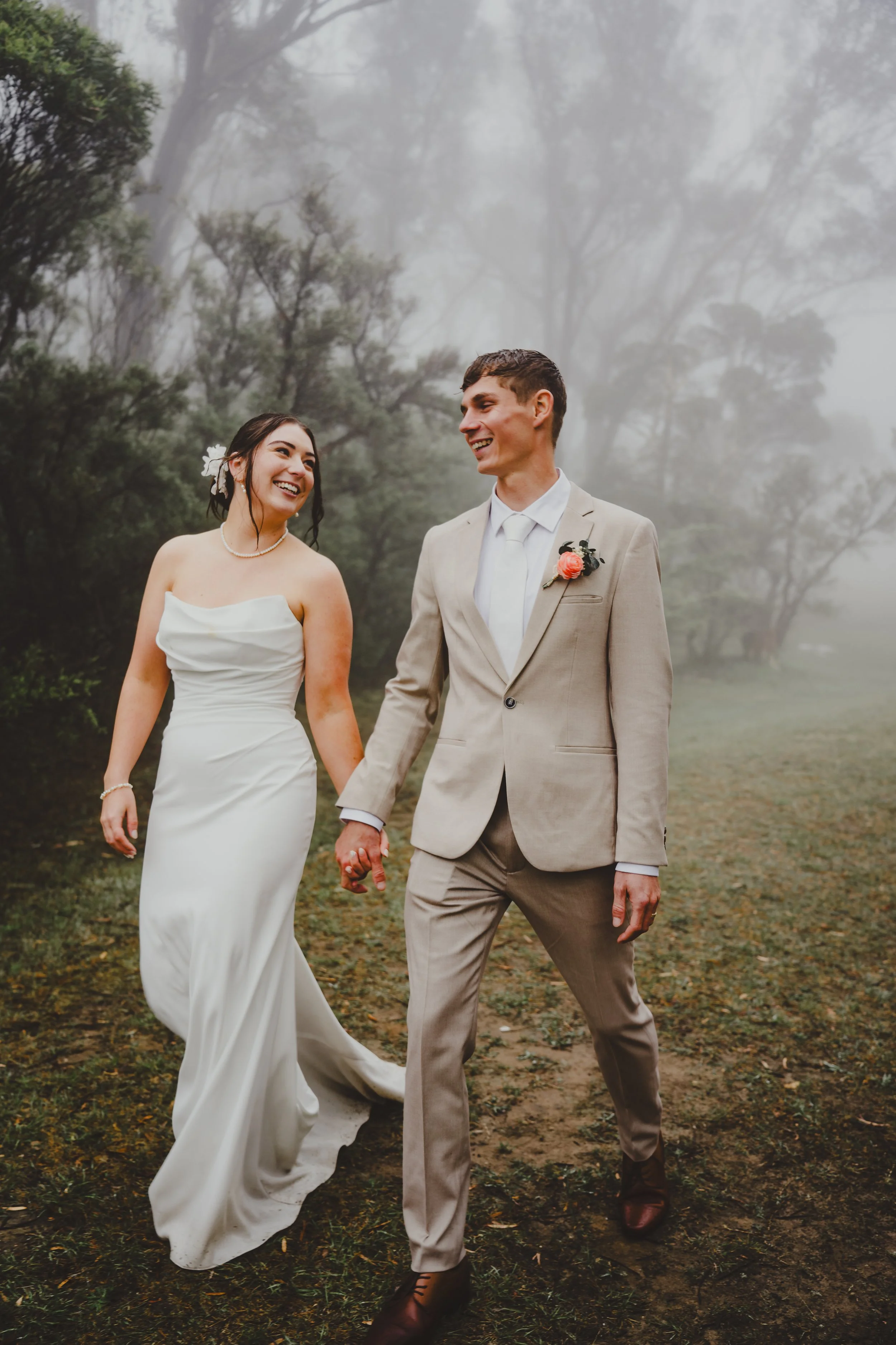 A bride and groom walking hand in hand outdoors on a foggy, wooded trail.