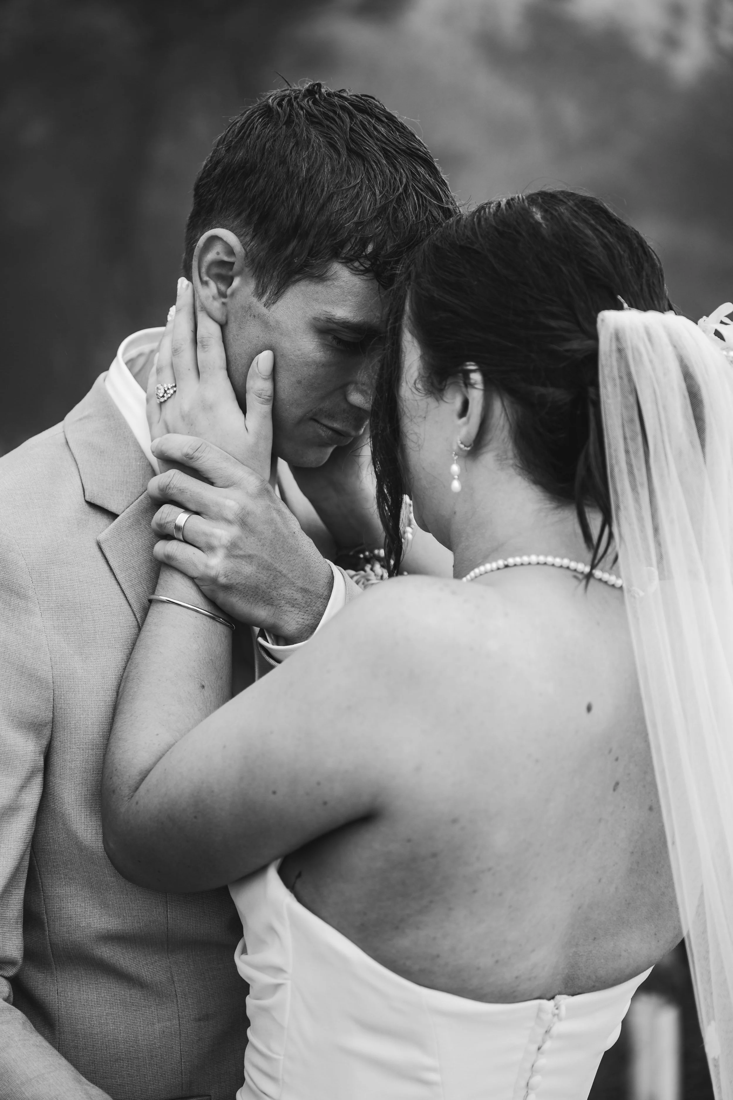A black-and-white photo of a bride and groom with their foreheads and noses touching, embracing outdoors.