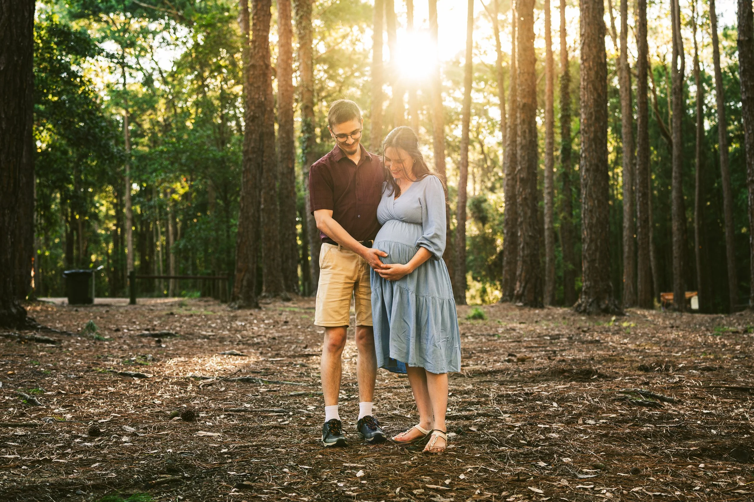 A pregnant woman in a light blue dress stands in a wooded park, holding her belly while a man in a dark shirt and beige shorts gently touches her belly. Sunlight filters through the trees.