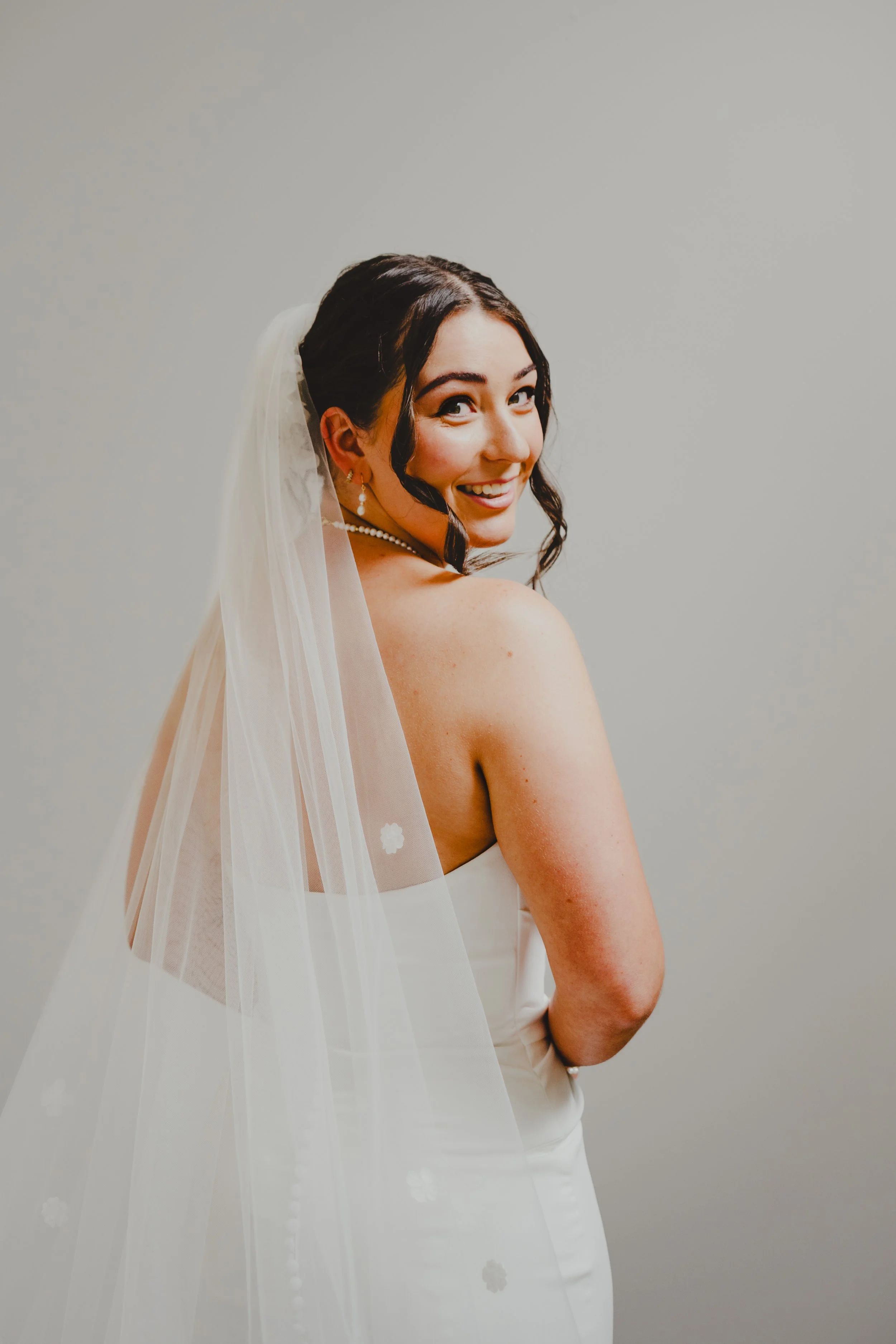 A bride in a white wedding dress with a veil and pearl jewelry, smiling and looking over her shoulder against a plain gray background.
