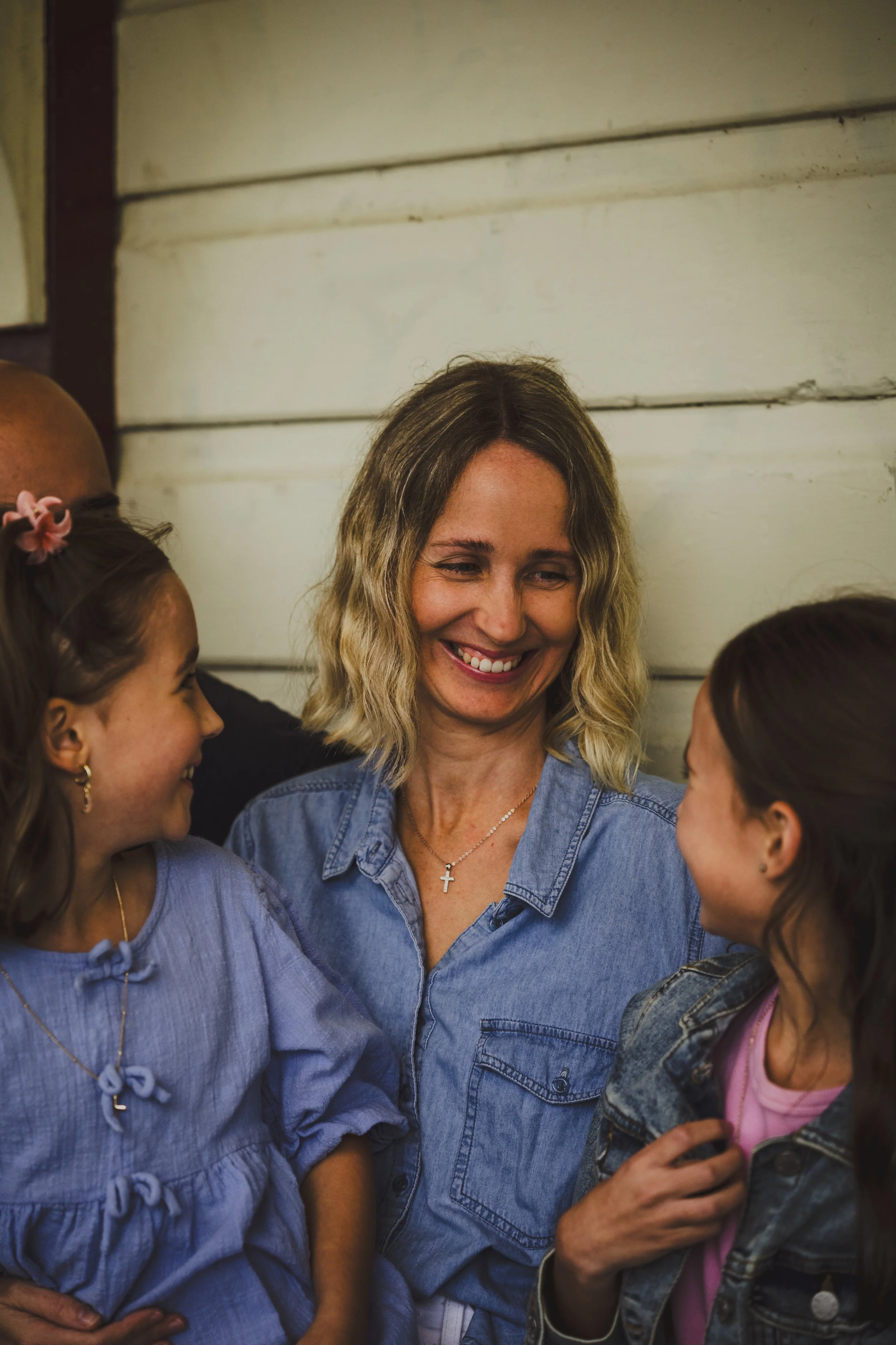 A woman smiling at two girls, one girl on each side, indoors against a wooden wall.