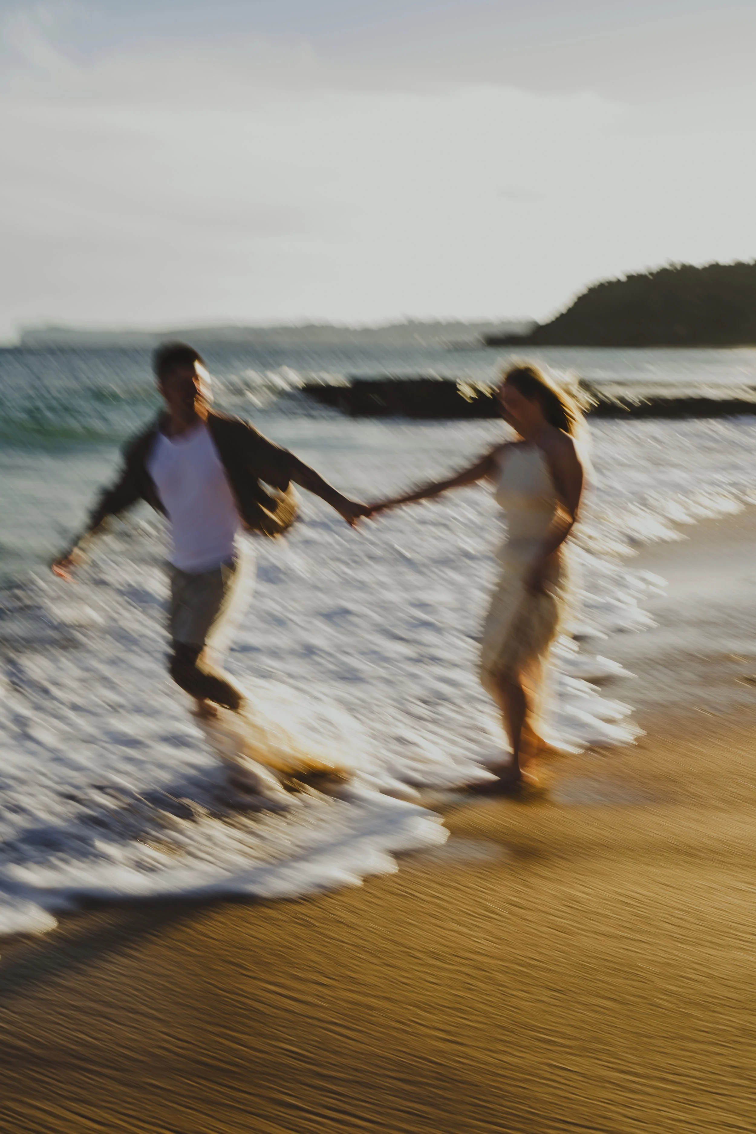 A blurred photo of a couple holding hands and playing in the ocean waves on a sandy beach during sunset.