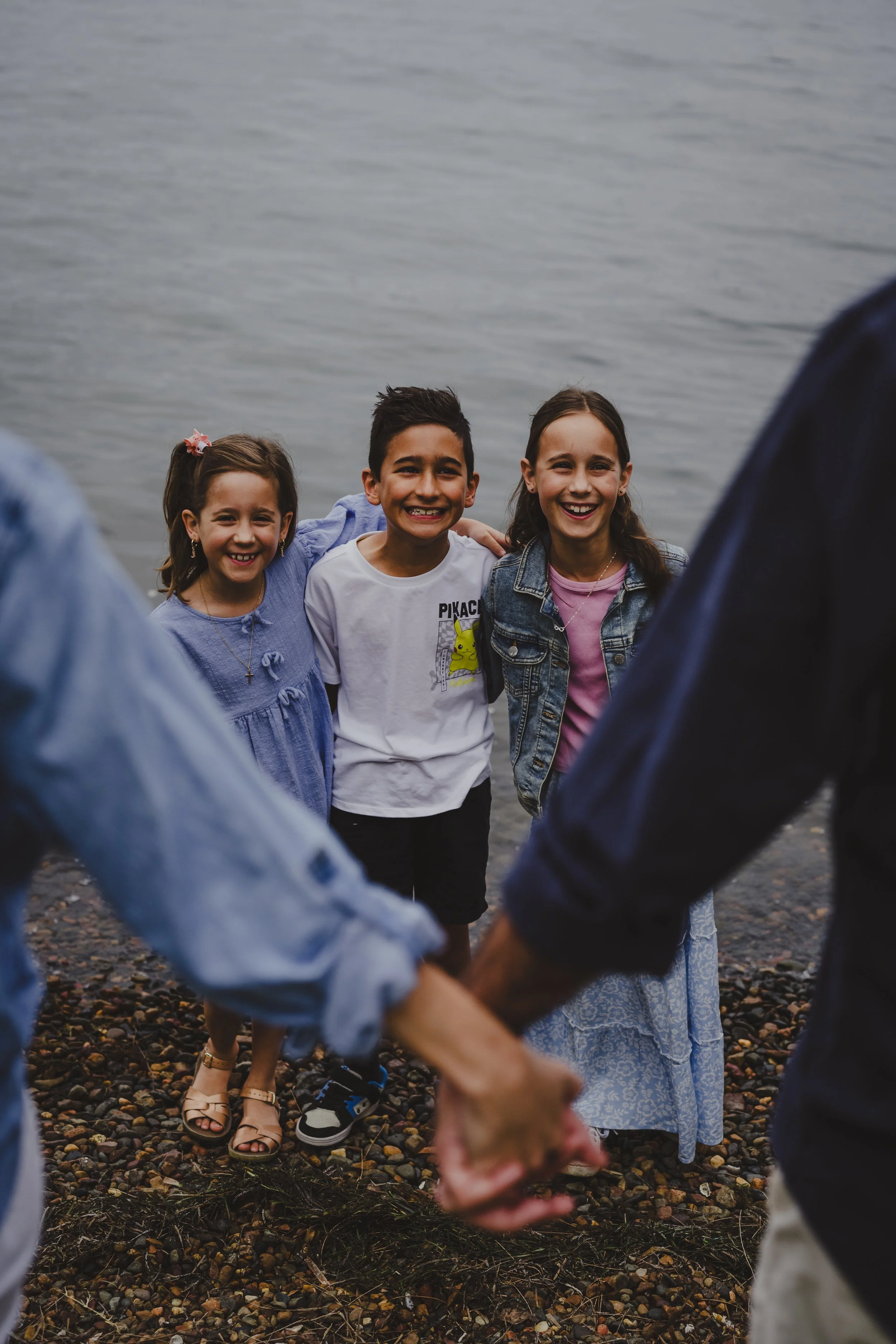 Three children standing outdoors near a body of water, smiling, with two people holding hands in the foreground.
