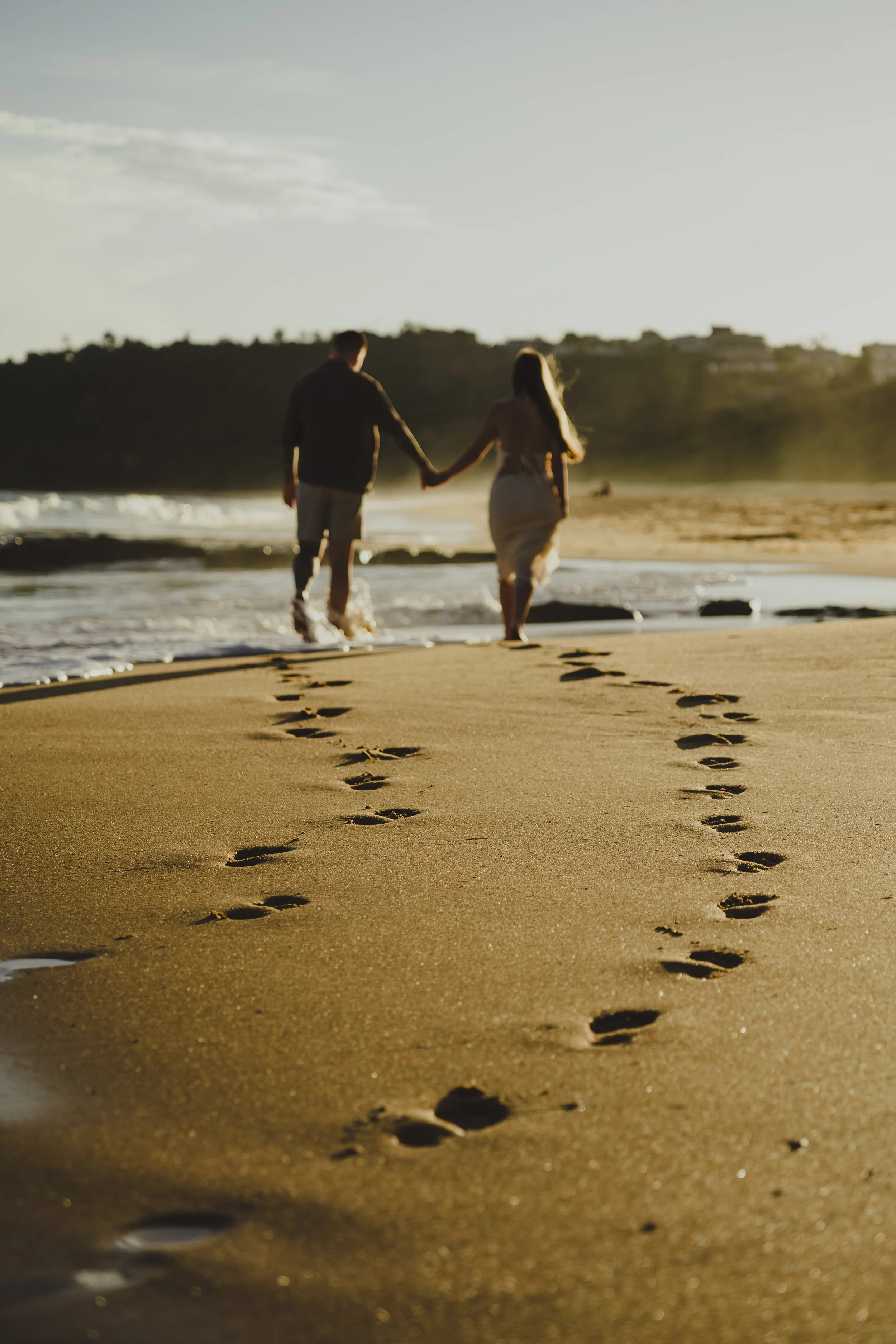 A couple holding hands and walking in the ocean at sunset, leaving footprints in the sand.