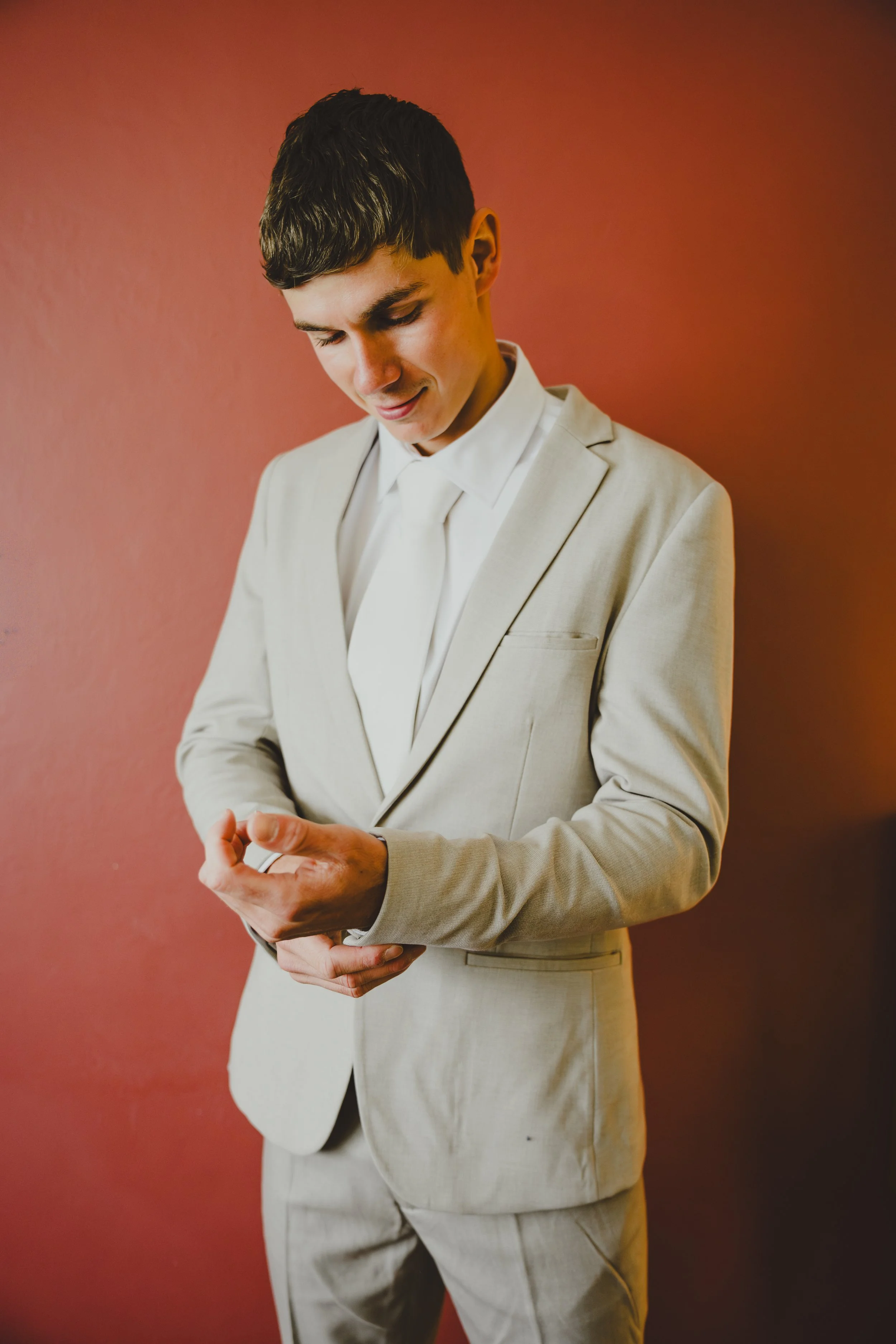 A young man in a light-colored suit and tie looking at his wristwatch against a reddish-brown background.