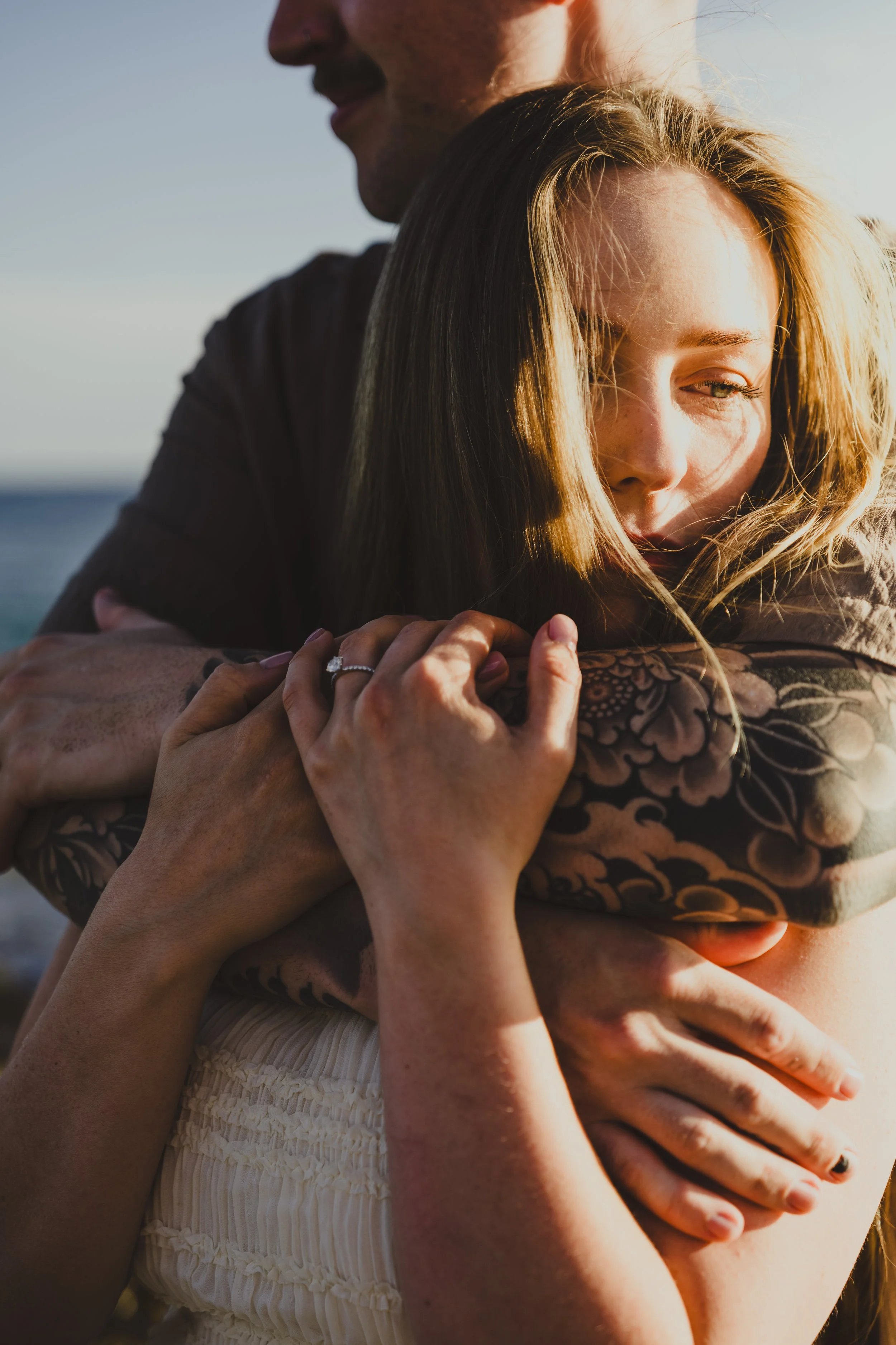 A man and woman embrace outdoors at sunset, with the woman resting her head on the man's shoulder. The woman has blonde hair and a ring on her finger; her arm has a floral tattoo. The man has dark hair and a mustache. The background shows a body of w