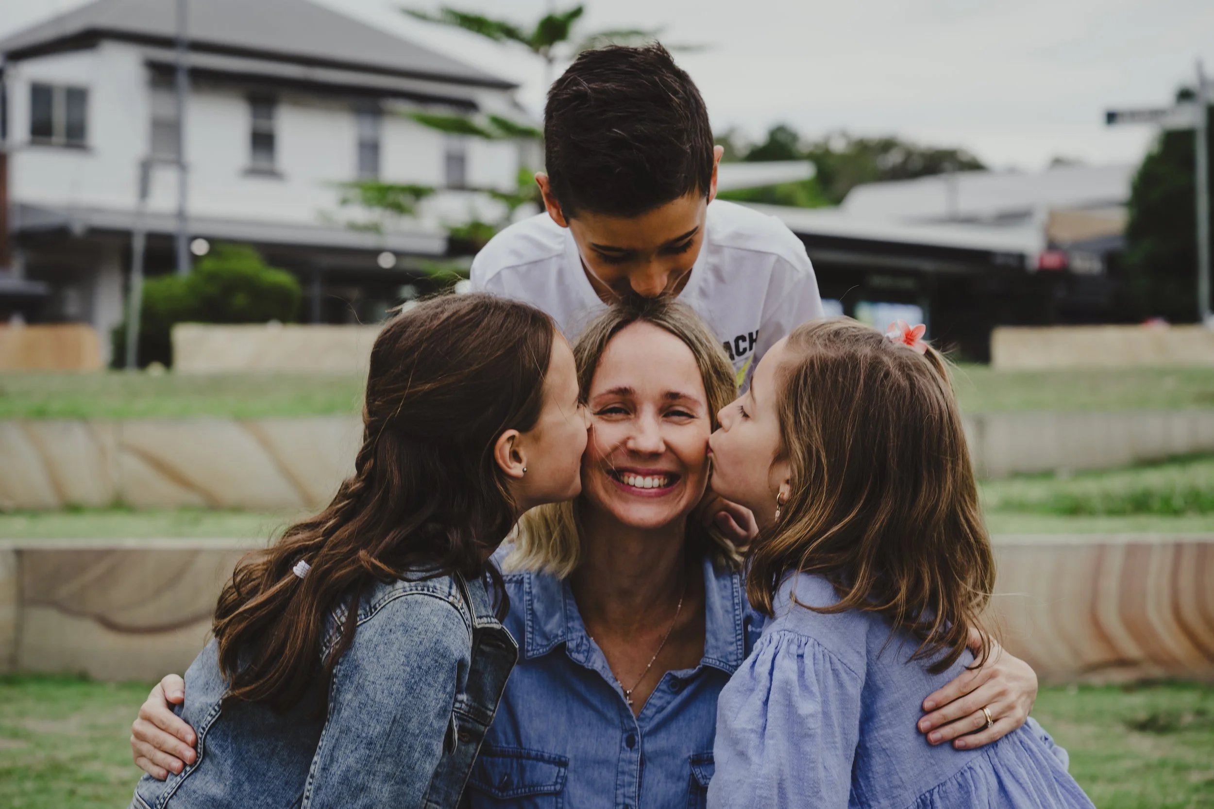A woman is smiling as she is kissed on both cheeks by two young girls, with a young boy leaning over her from behind, all outdoors in a park or garden area.