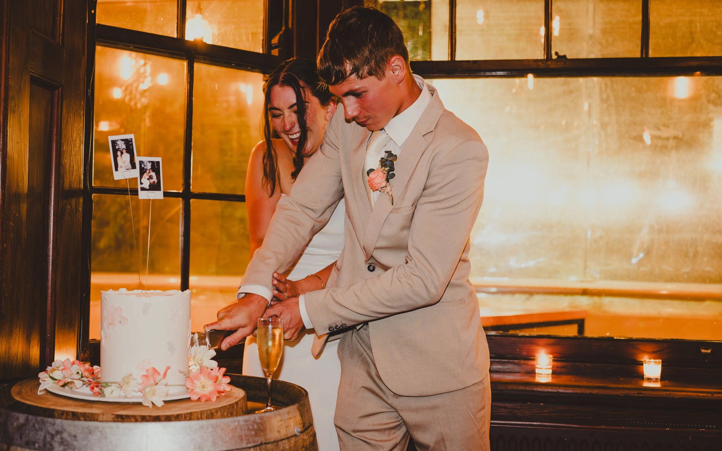 A bride and groom cutting their wedding cake together, smiling and celebrating at their wedding reception with a champagne flute nearby, decorated with flowers and photographs, in warm indoor lighting.