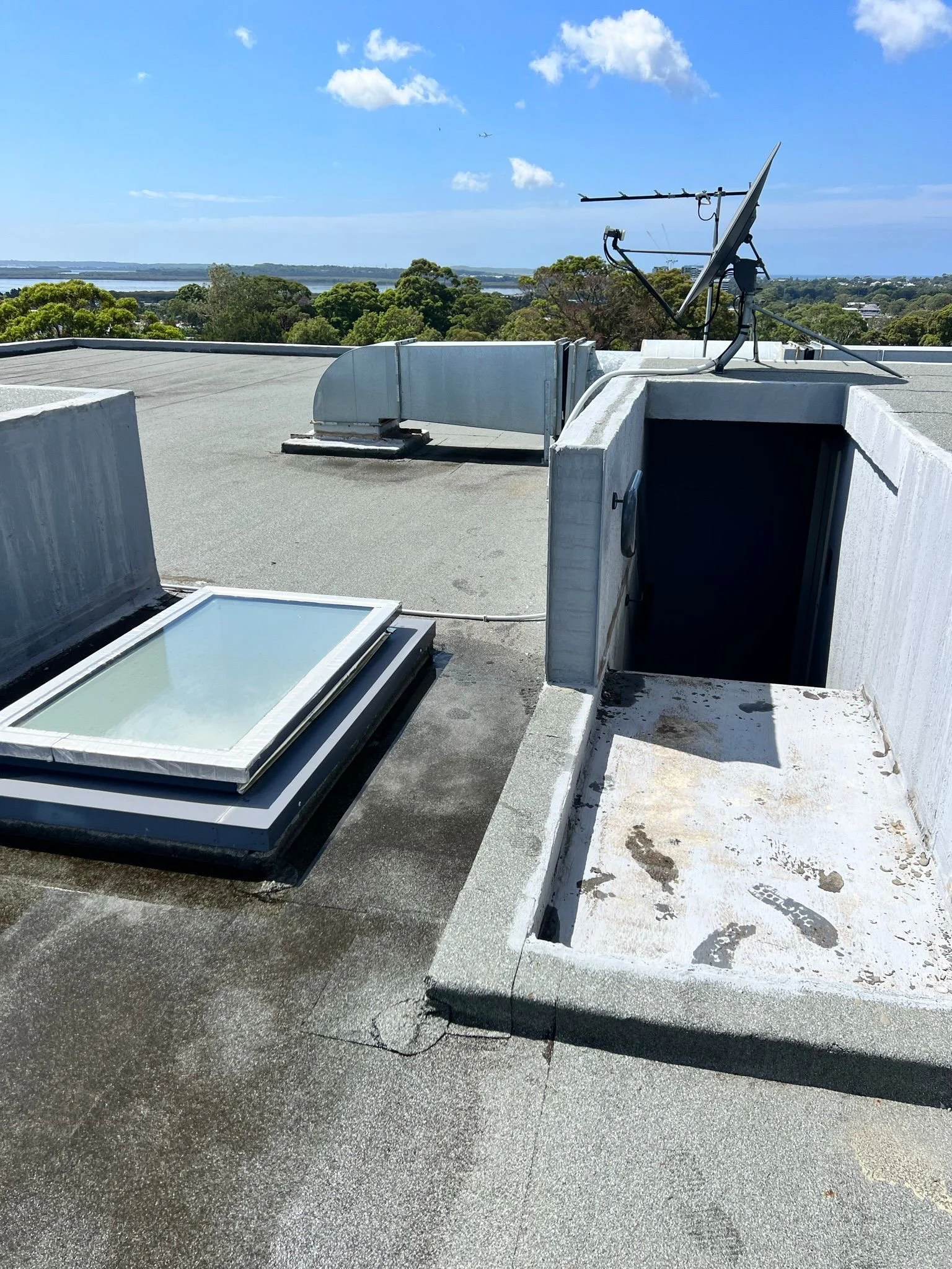 View of a rooftop with skylight window, metal vent, satellite dish, and an open hatch, overlooking trees and water in the distance under a blue sky with scattered clouds.