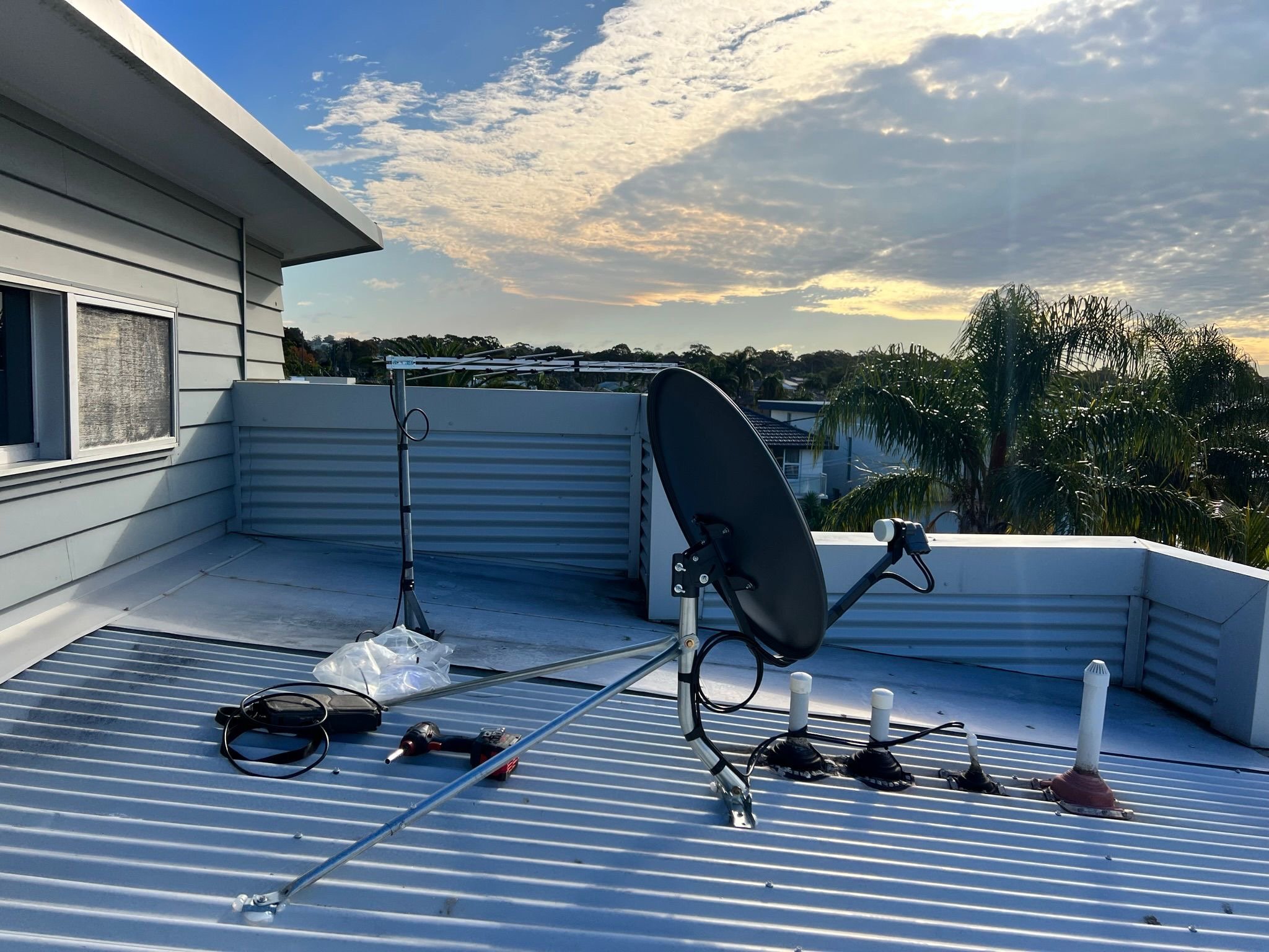 Rooftop with satellite dish, antenna, and various installation tools during sunset, with a house window and palm trees in the background.