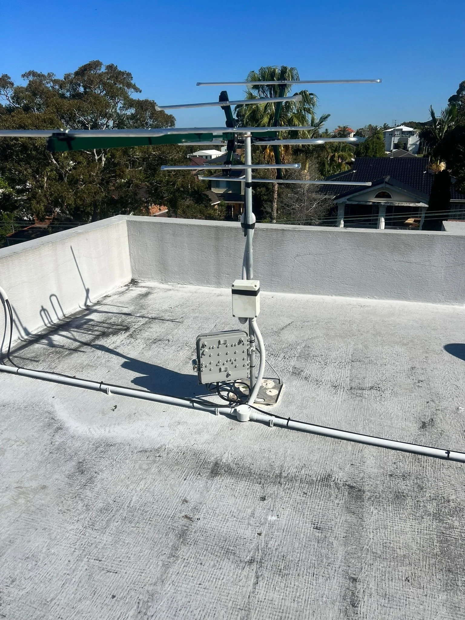 A rooftop with a television antenna and a communication box, with trees and houses in the background under a clear blue sky.