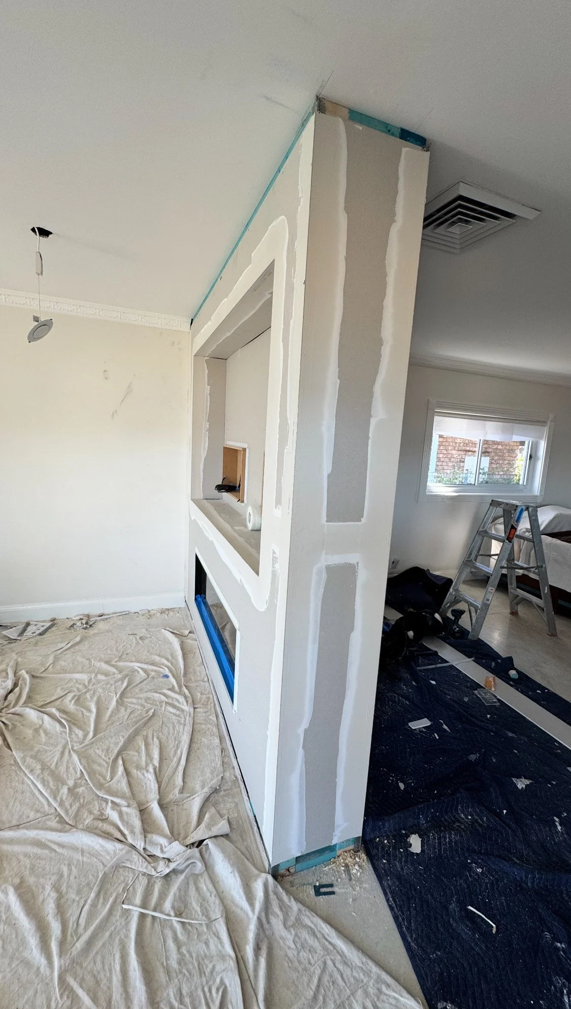 Interior view of a room under construction or renovation, with a partial wall with drywall and insulation visible, a ladder, and construction materials on the floor.