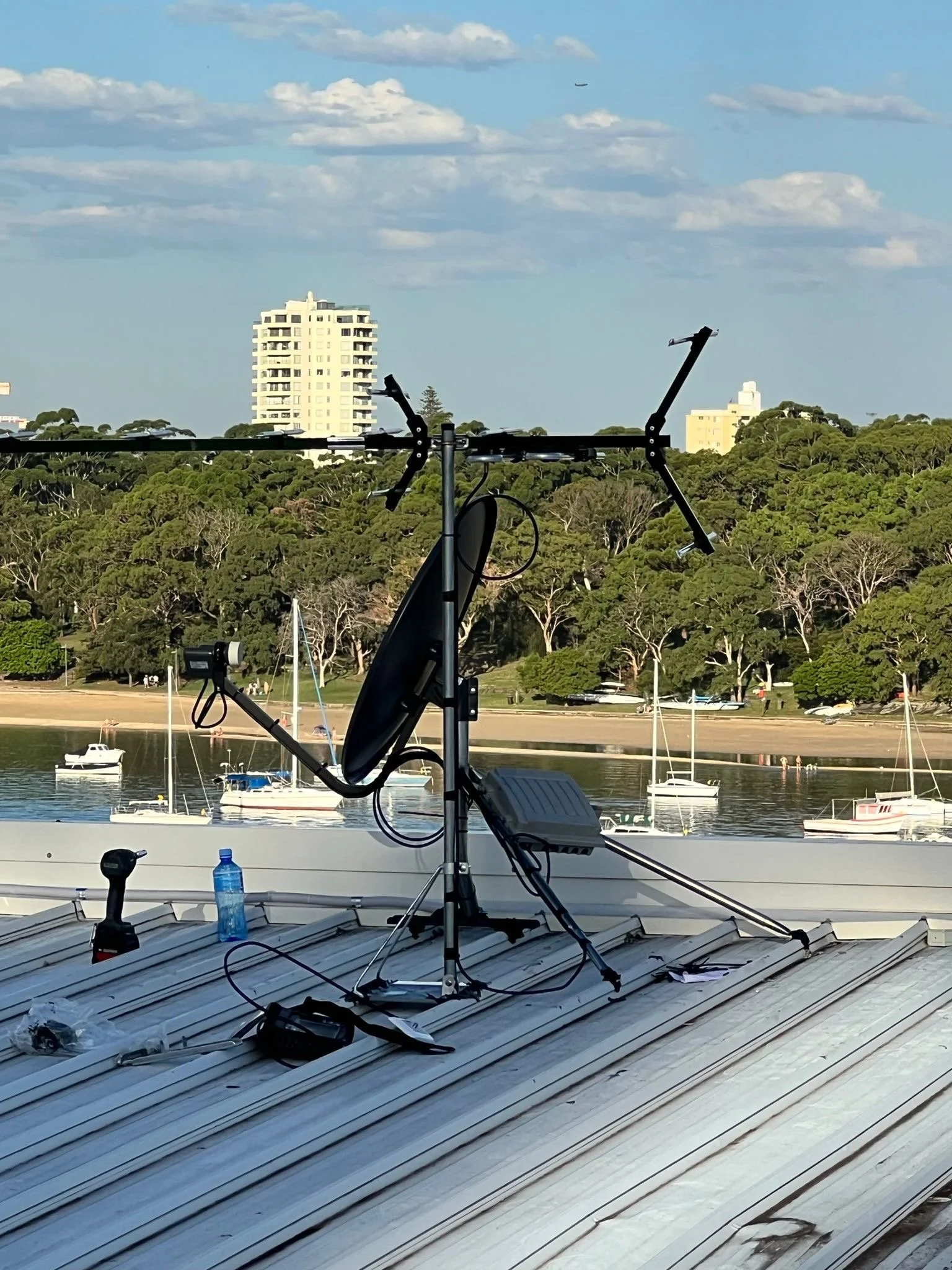 A rooftop with a TV antenna and a satellite dish, overlooking a river with boats and a park with trees and buildings in the background.