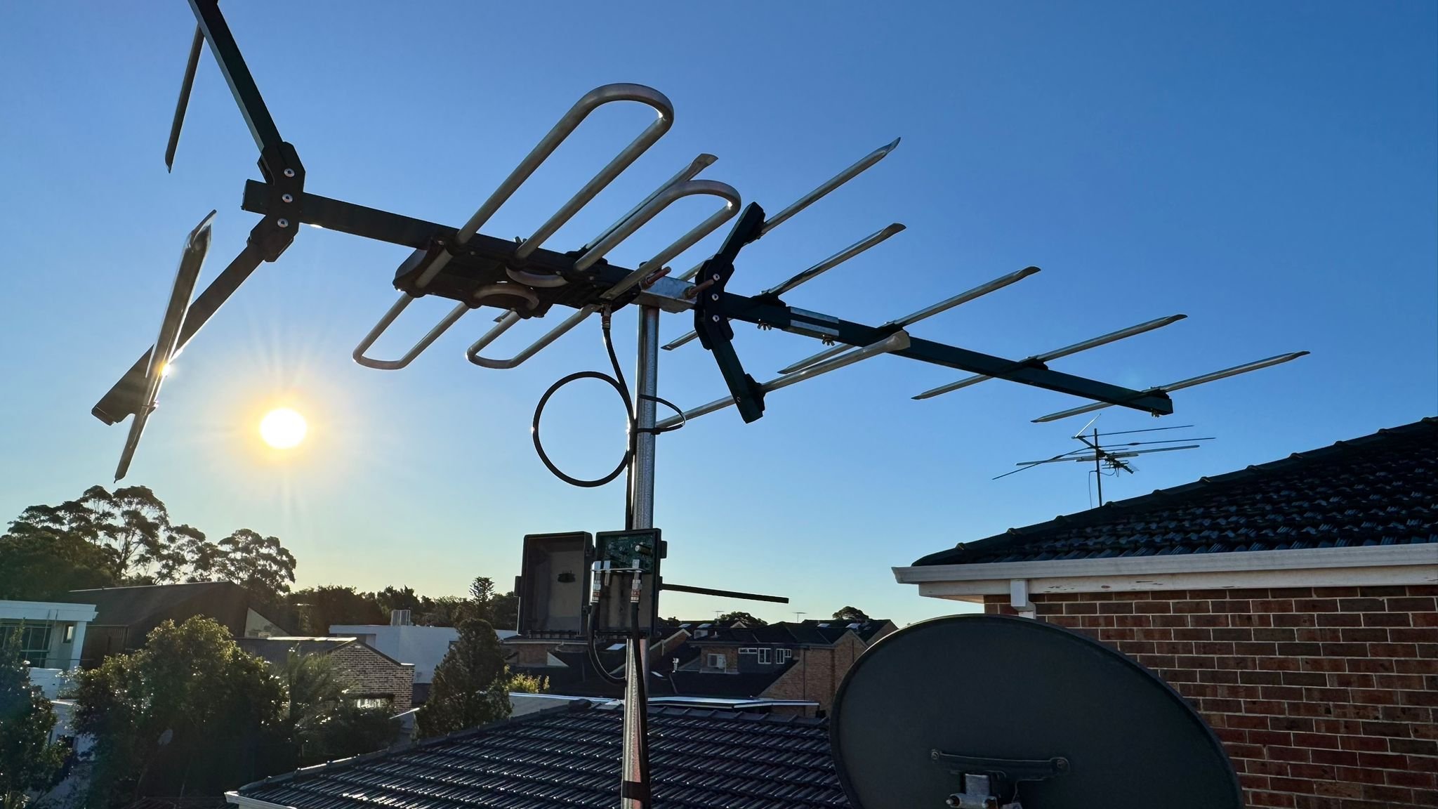 A TV antenna and satellite dish mounted on a rooftop against a clear blue sky with the sun shining.