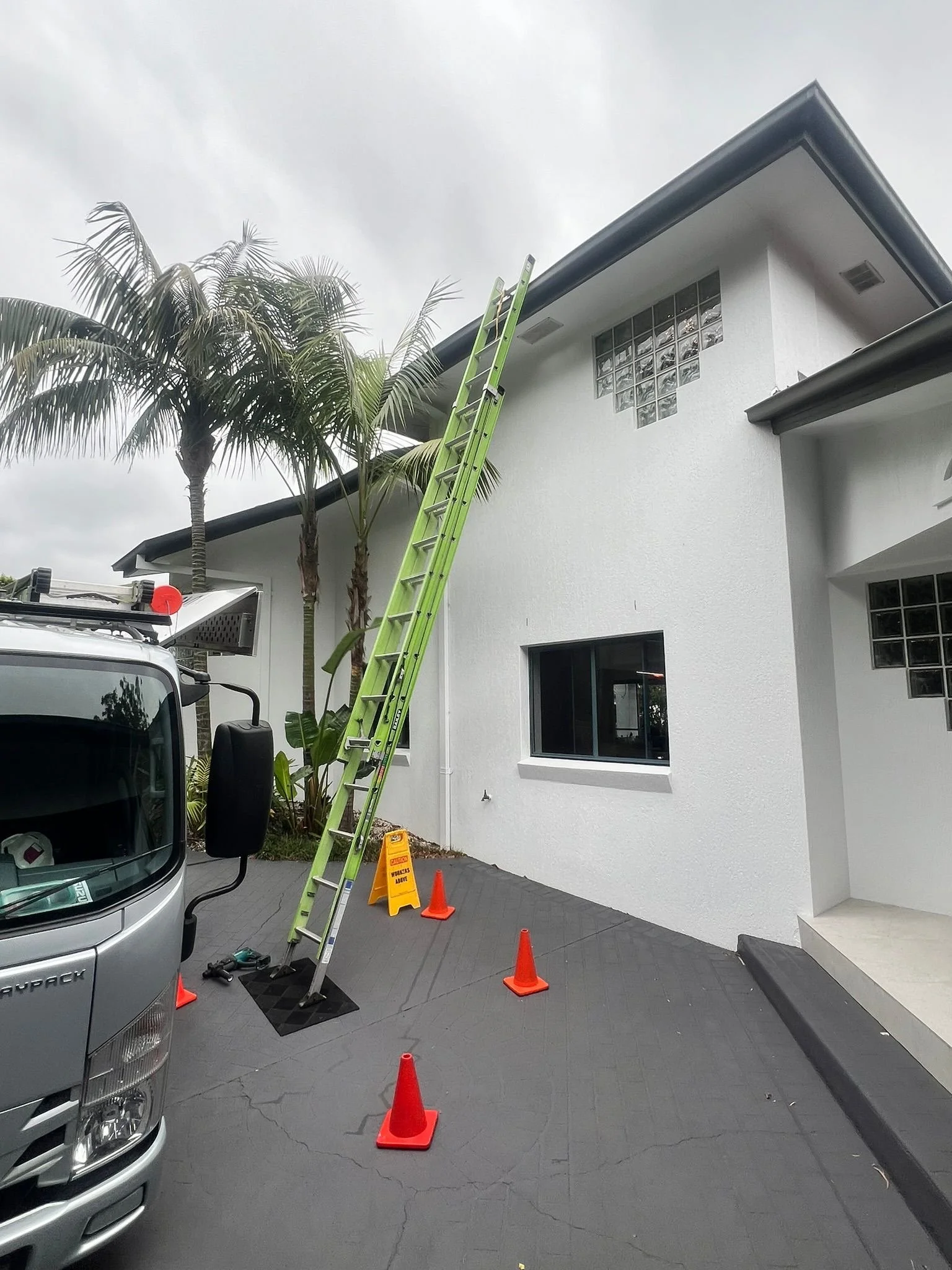 A residential building with a tall ladder leaning against the wall near a palm tree, surrounded by orange traffic cones and a yellow wet floor sign on the driveway.