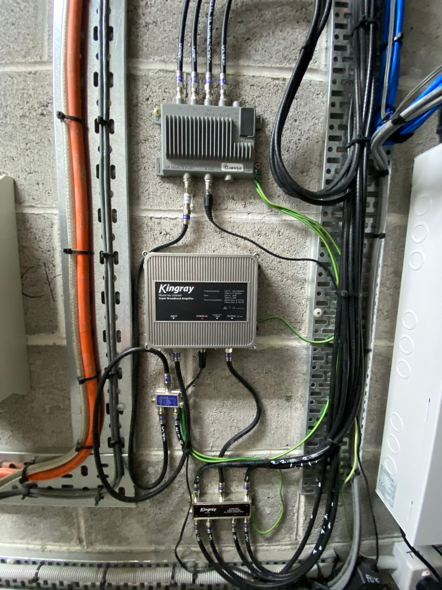 Electrical equipment and wiring mounted on a concrete wall, including a Kingray broadband amplifier, a small gray electronic device, and various cables and connectors.