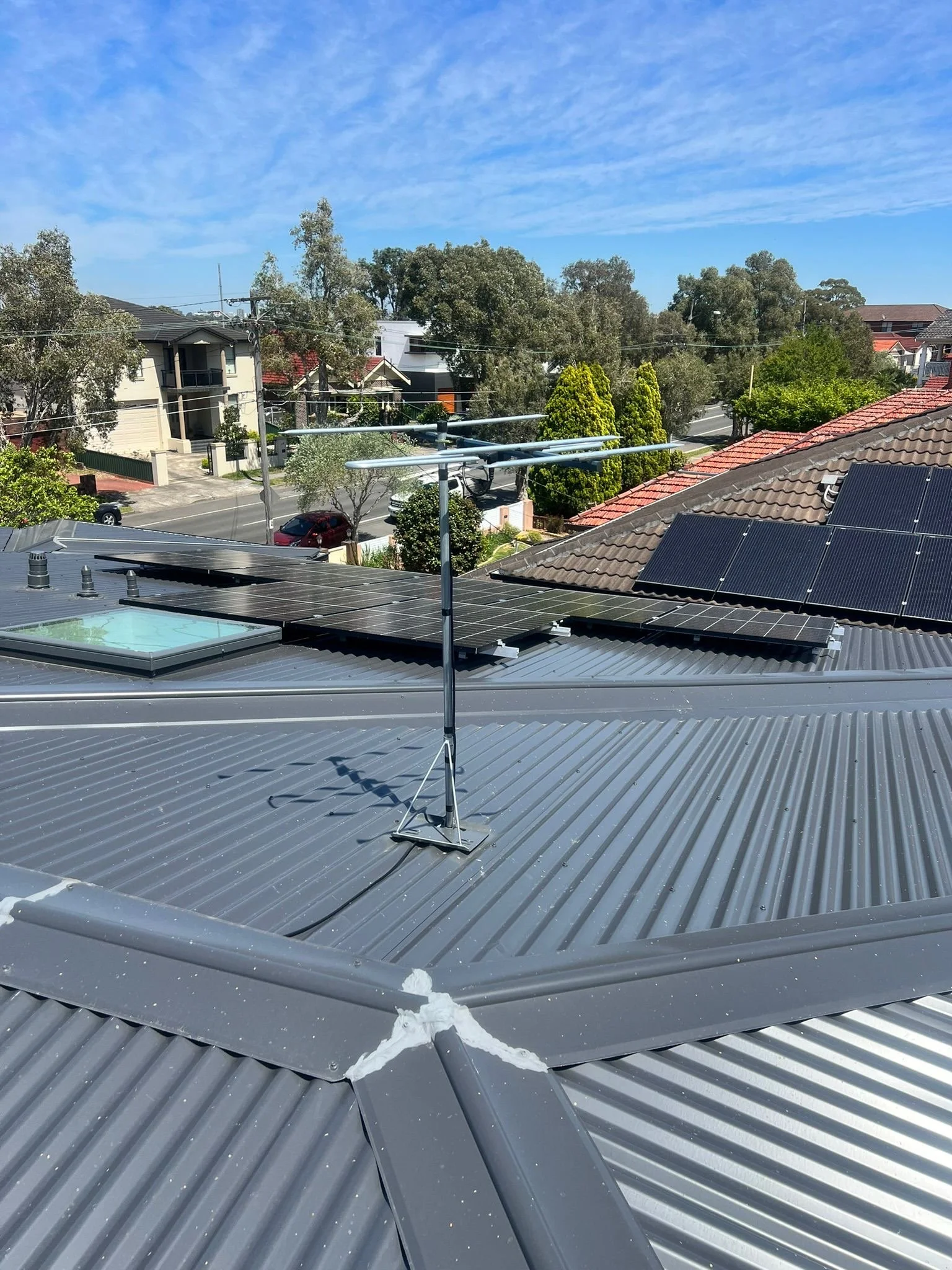 View of a metal rooftop with solar panels and a TV antenna mounted on a pole, with a suburban neighborhood and trees in the background under a partly cloudy blue sky.