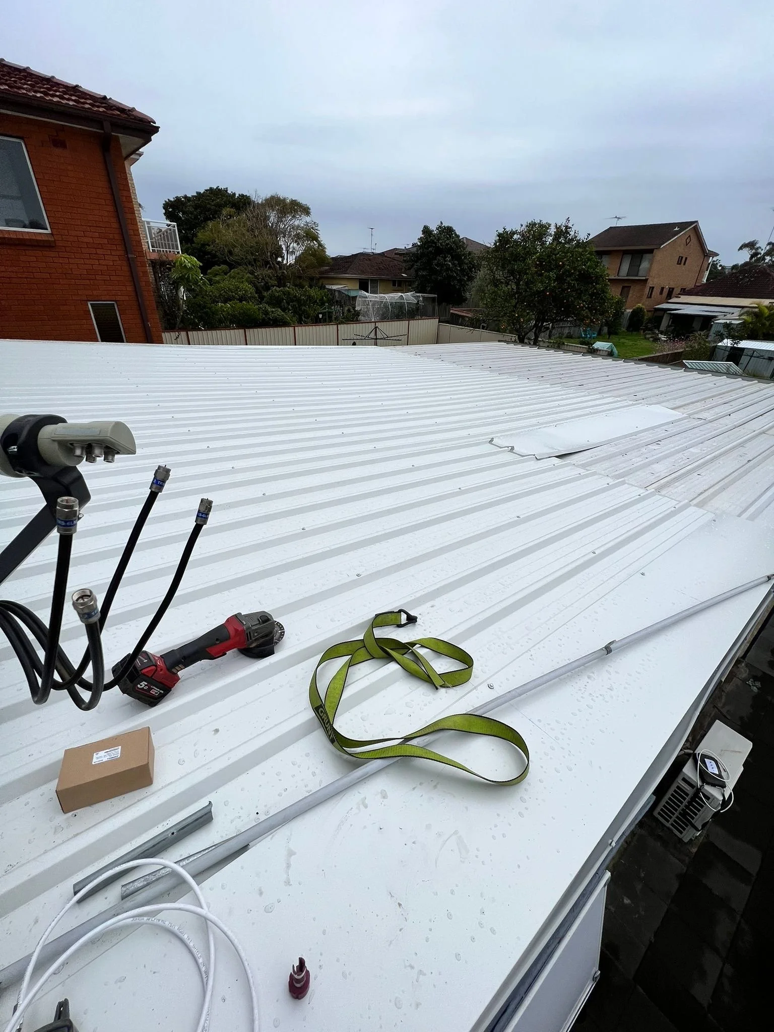 View of a white metal roofing with tools and equipment on top, including a cordless drill, a green strap, a cardboard box, and cables, with residential houses and trees in the background.