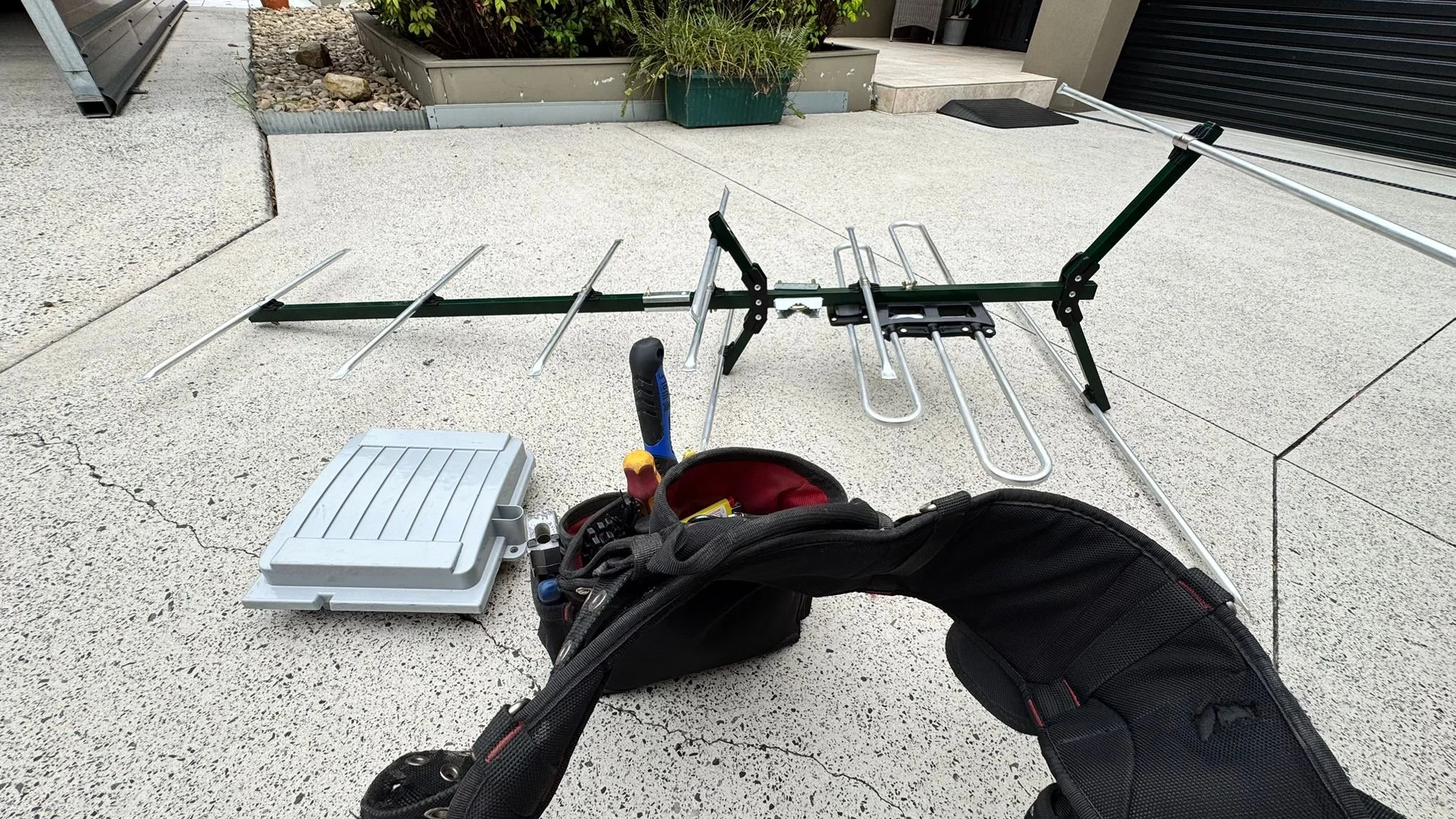 Yagi antenna, a toolbox, and a black backpack with tools are set up on a concrete patio near a garden bed.