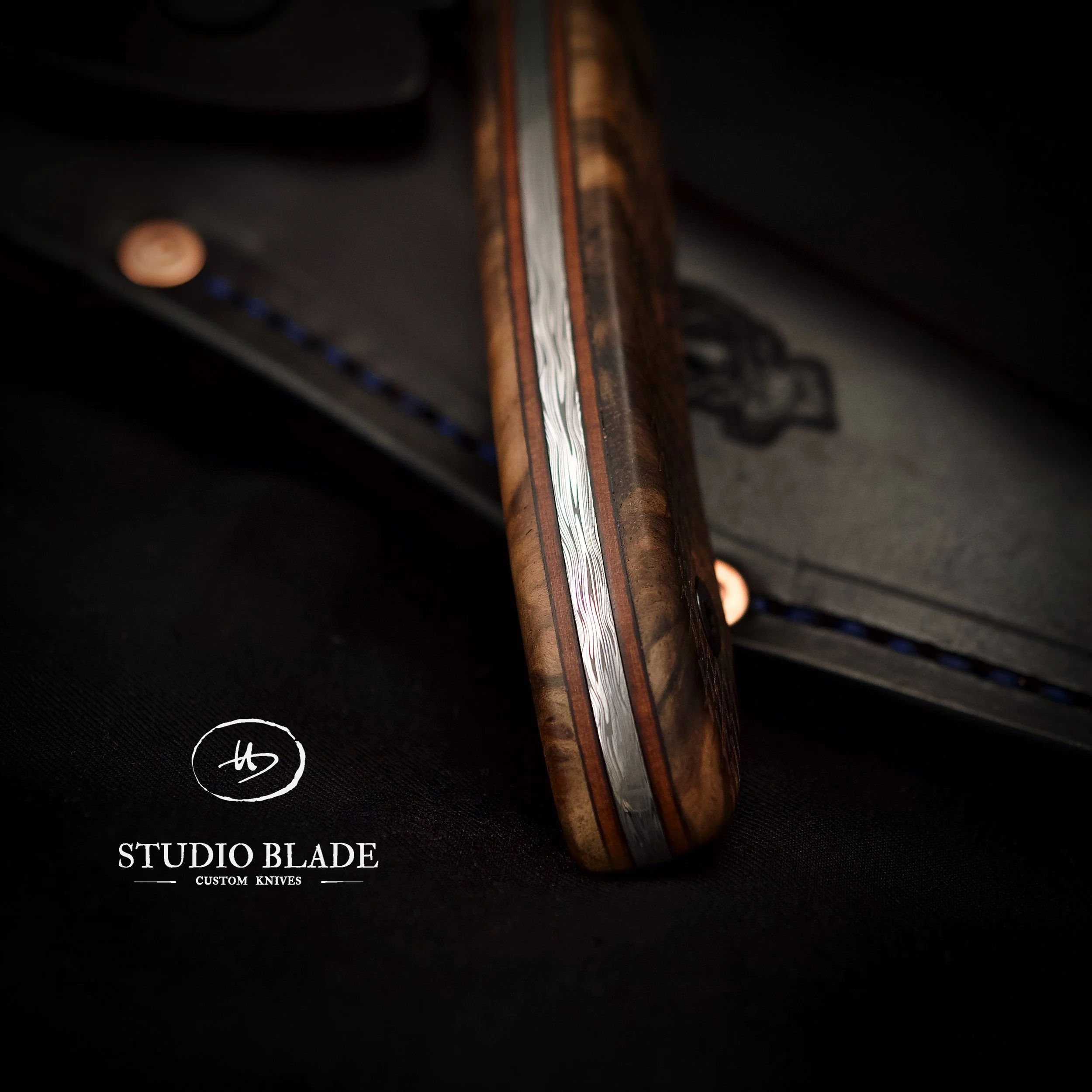 Close-up of a custom Turkish Walnut burl knife handle with damascus metal accents on a black surface, with the logo and text 'Studio Blade Custom Knives' in the lower left corner.