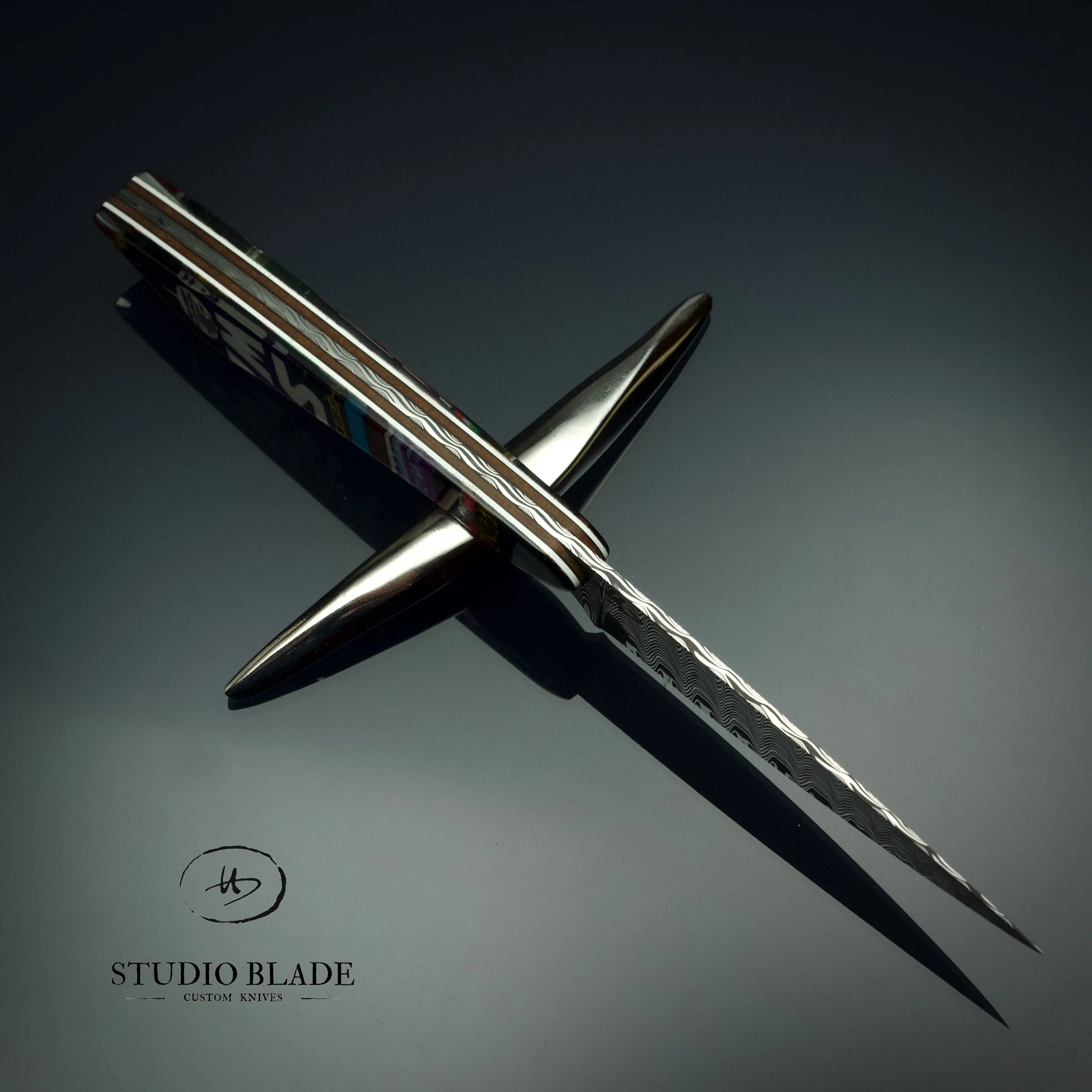 A modern, artistic kitchen and table knife with a decorative patterned blade and a polished metal handle, branded with 'STUDIO BLADE'. It is positioned diagonally on a gray surface.