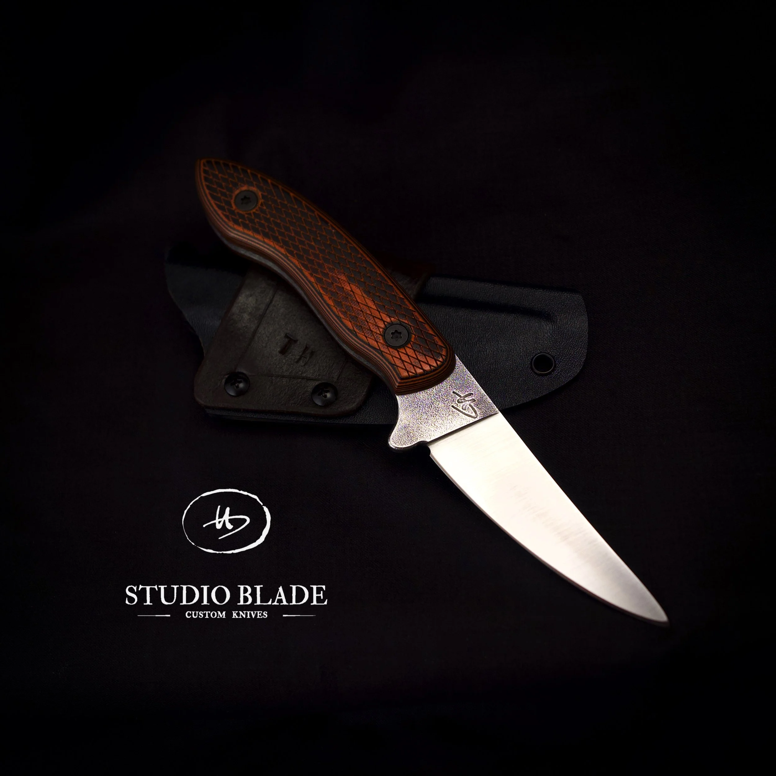A custom-made hunting knife by Studio Blade with a textured blade engraved with a logo and orange G10 handles with a diamond pattern, resting on a black surface. With a logo that reads 'Studio Blade Custom Knives'.
