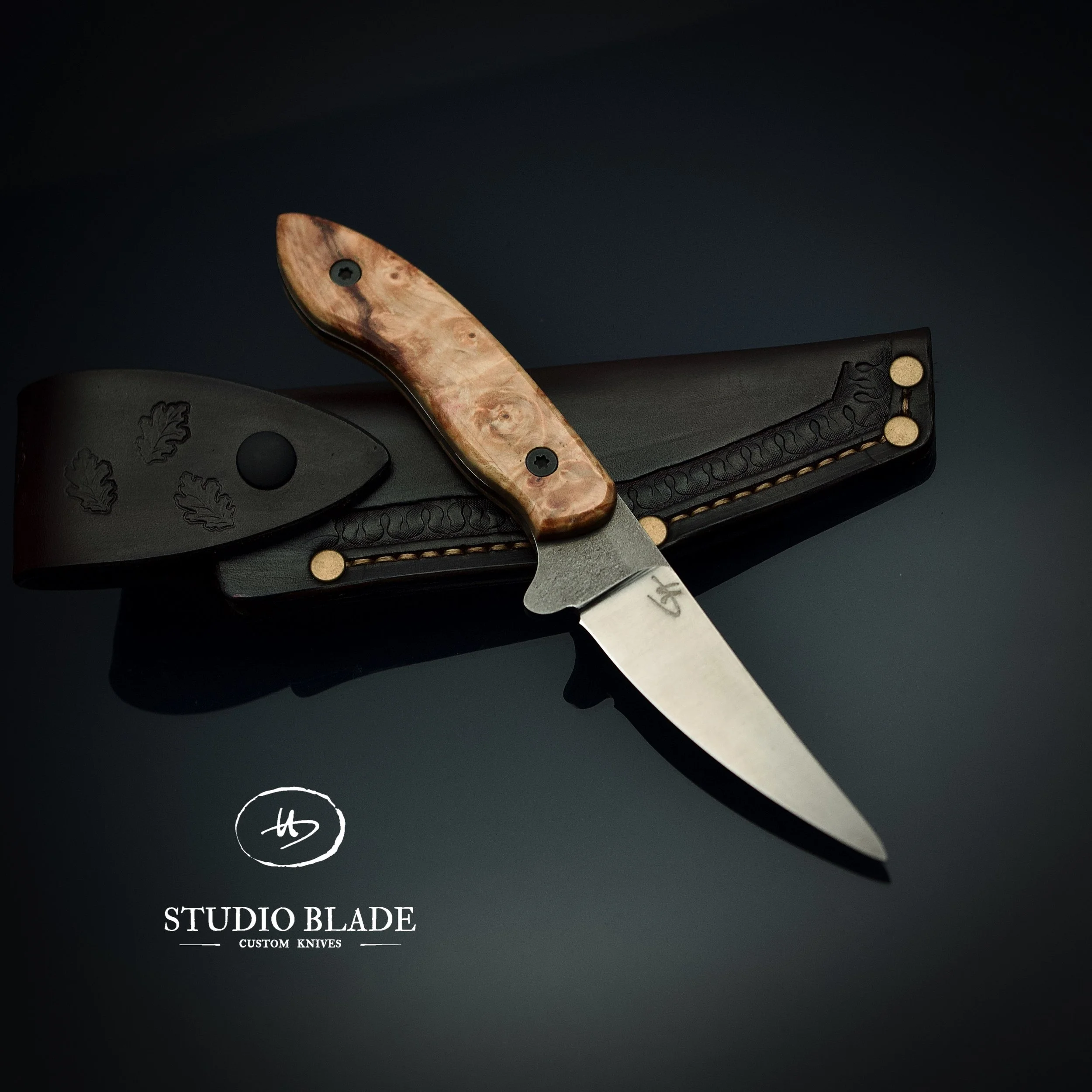 A custom hunting knife with a Maple handle and stainless steel blade, resting on a handmade black leather sheath with an embossed oak leaf and copper rivets for reinforcement. The image features the logo of Studio Blade, a custom knife maker.