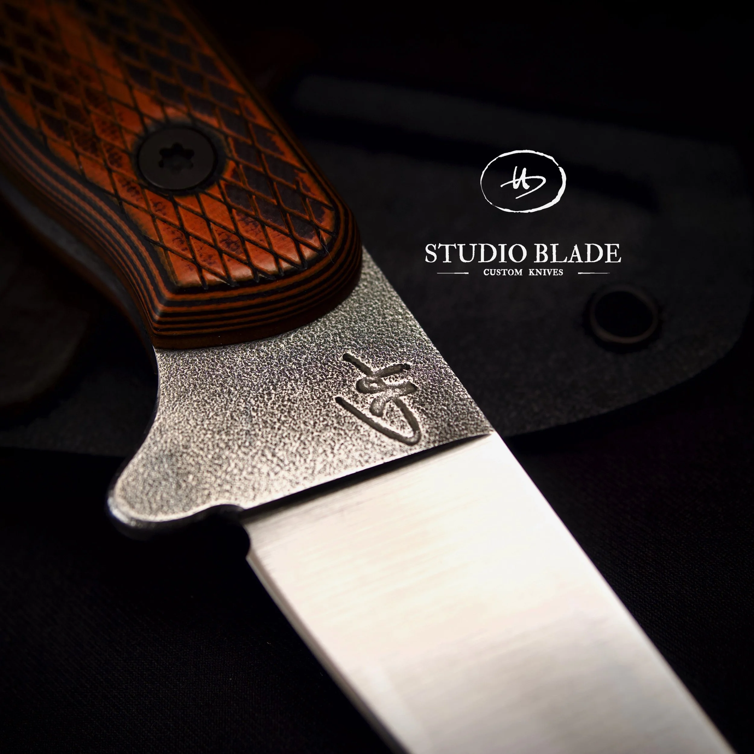 Close-up of a custom-made knife by Studio Blade with a textured blade engraved with a logo, and an orange G10 handle with a diamond pattern, resting on a black surface.