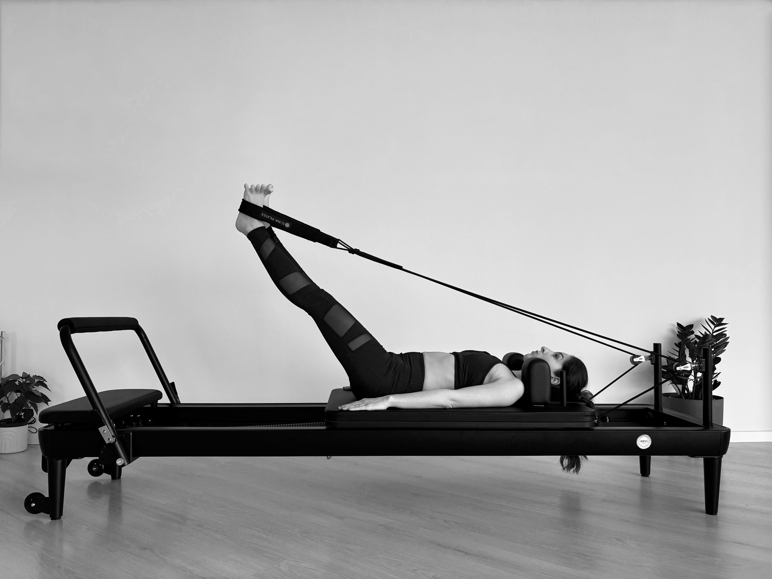 A woman lying on a Pilates reformer machine while doing a leg exercise with resistance bands. The room has a plain white wall, wooden floor, and potted plants in the background.