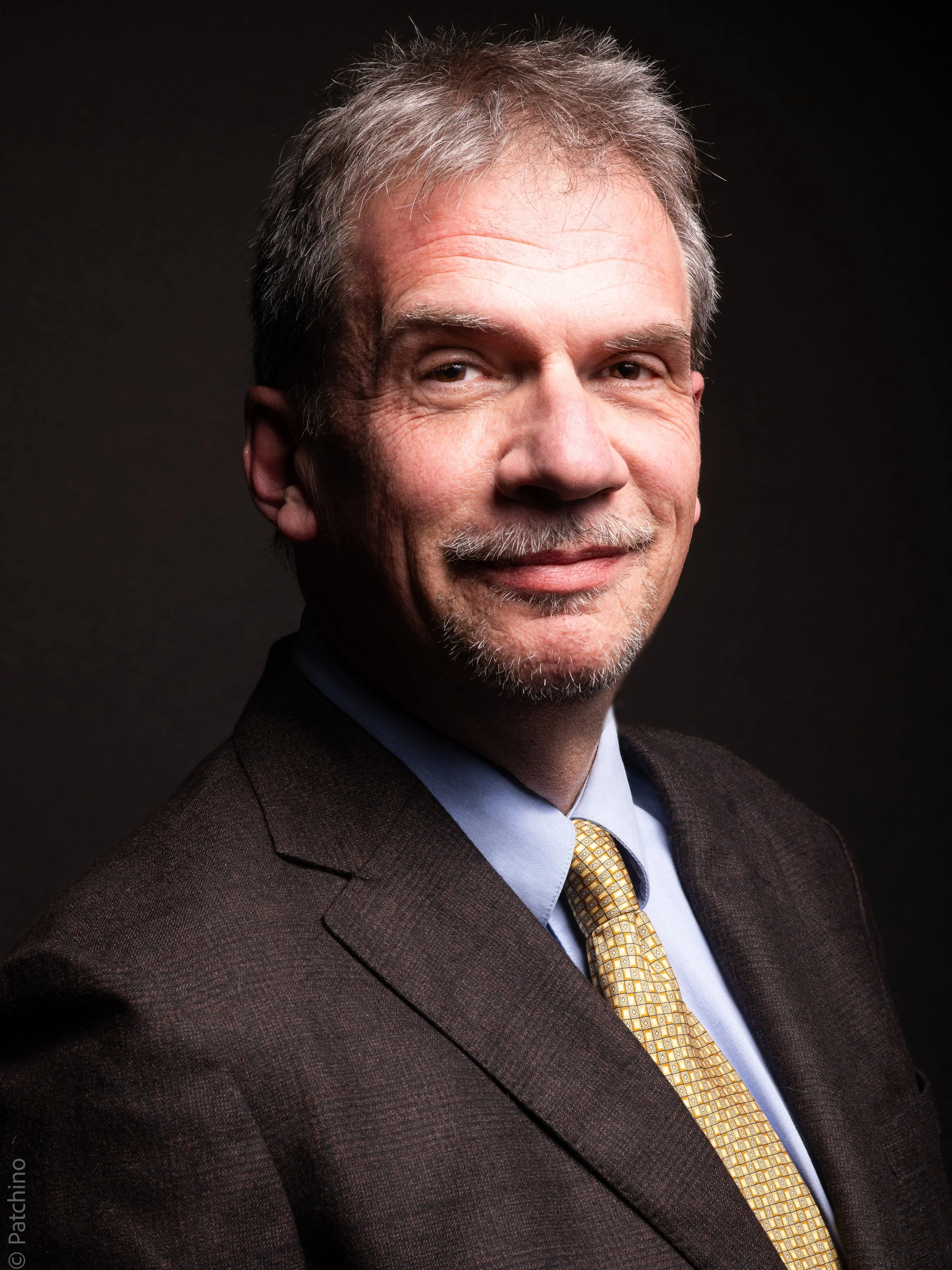 Pierre Diot, portant une chemise bleue, une cravate jaune et un costume foncé, souriant légèrement, contre un fond noir.